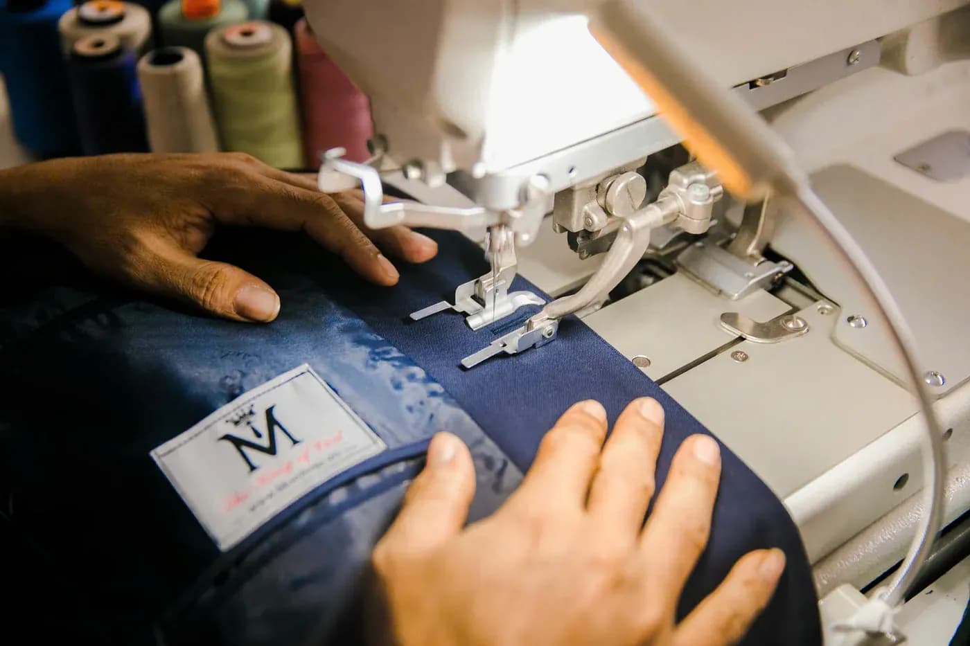 Bàn tay thợ may đưa thân vest xanh navy qua máy, nhãn Mon Amie đã gắn
