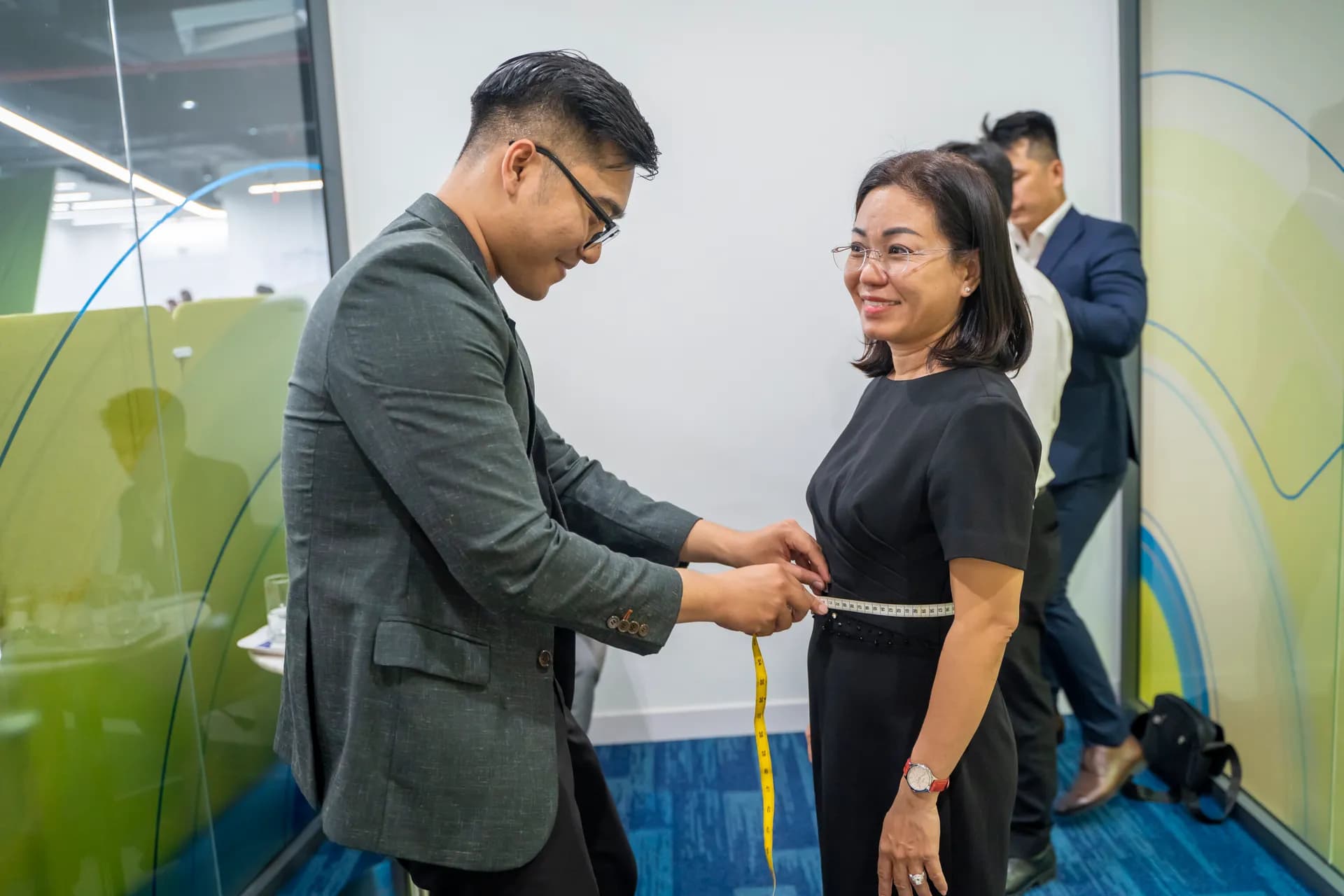 Khách nữ đầm đen mỉm cười khi thợ may đo vòng eo bằng thước dây vàng