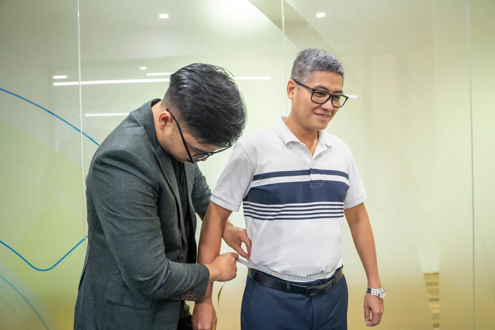 Khách nam áo polo sọc mỉm cười khi thợ may vest xám đo bắp tay