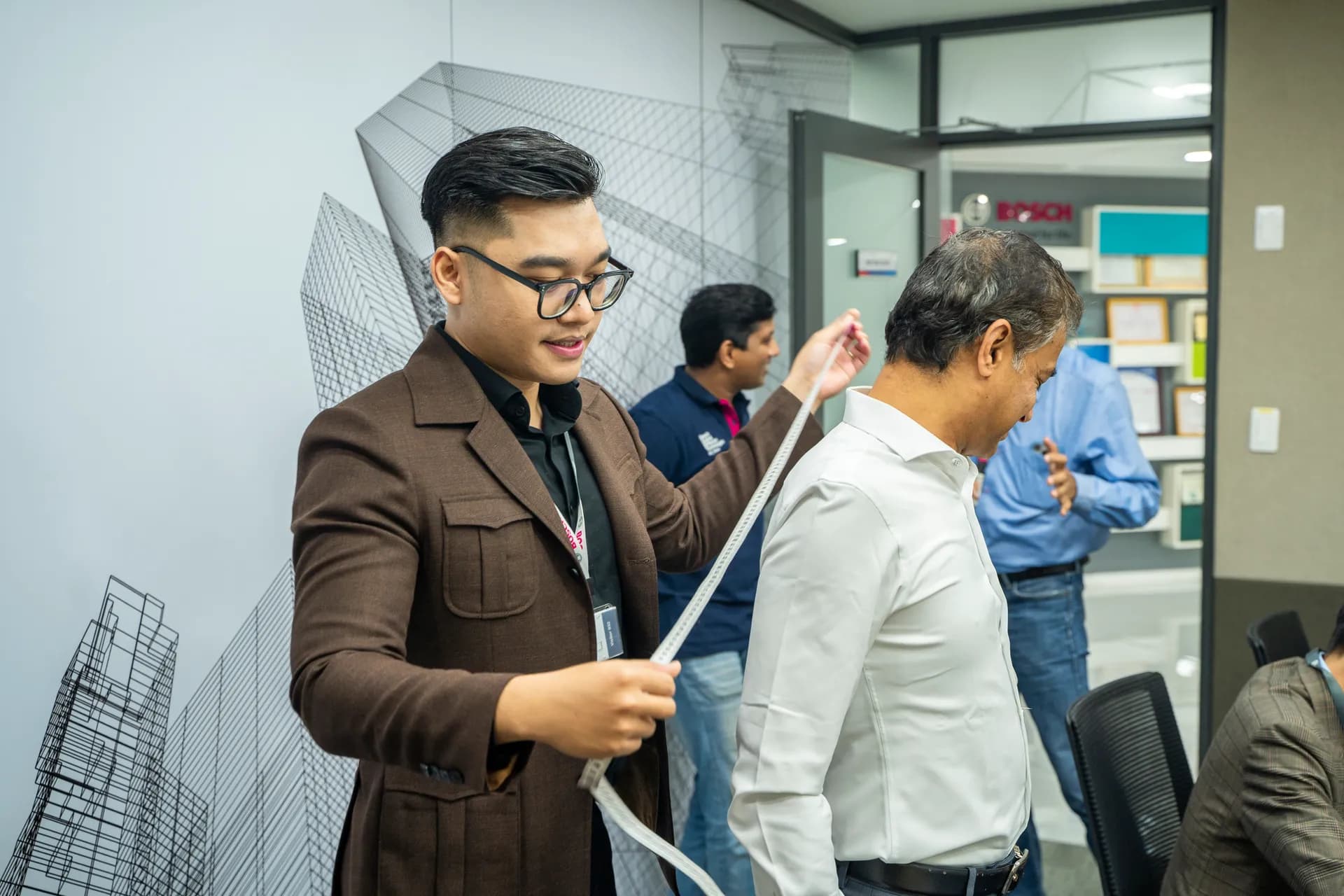 Thợ mặc áo khoác nâu kéo thước dây đo thân cho khách sơ mi trắng, nụ cười nhẹ