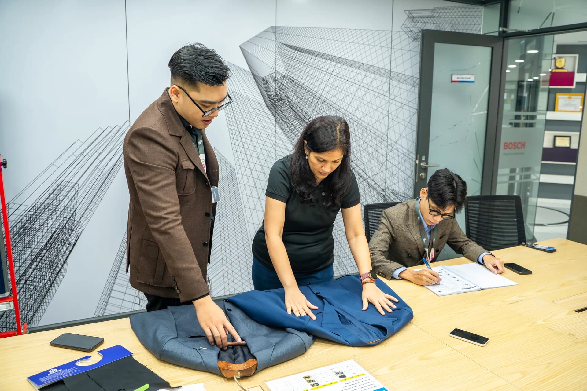 Bàn tư vấn trải áo vest xanh navy và sổ số đo, thợ áo khoác nâu cùng khách nữ