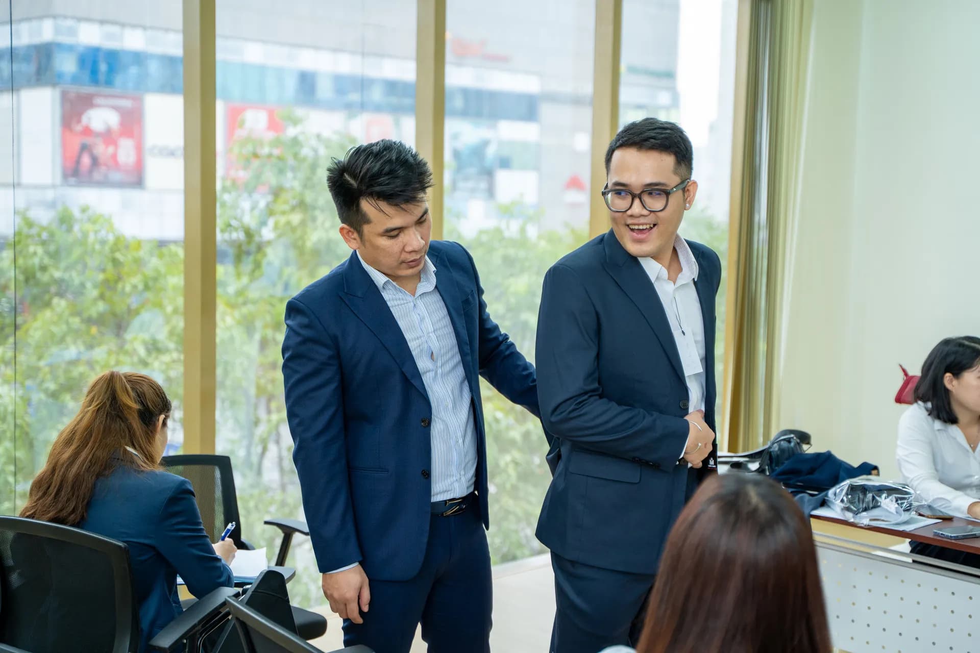 Hai nam nhân viên mặc suit navy thử đồng phục mới bên cửa kính, cười tự nhiên