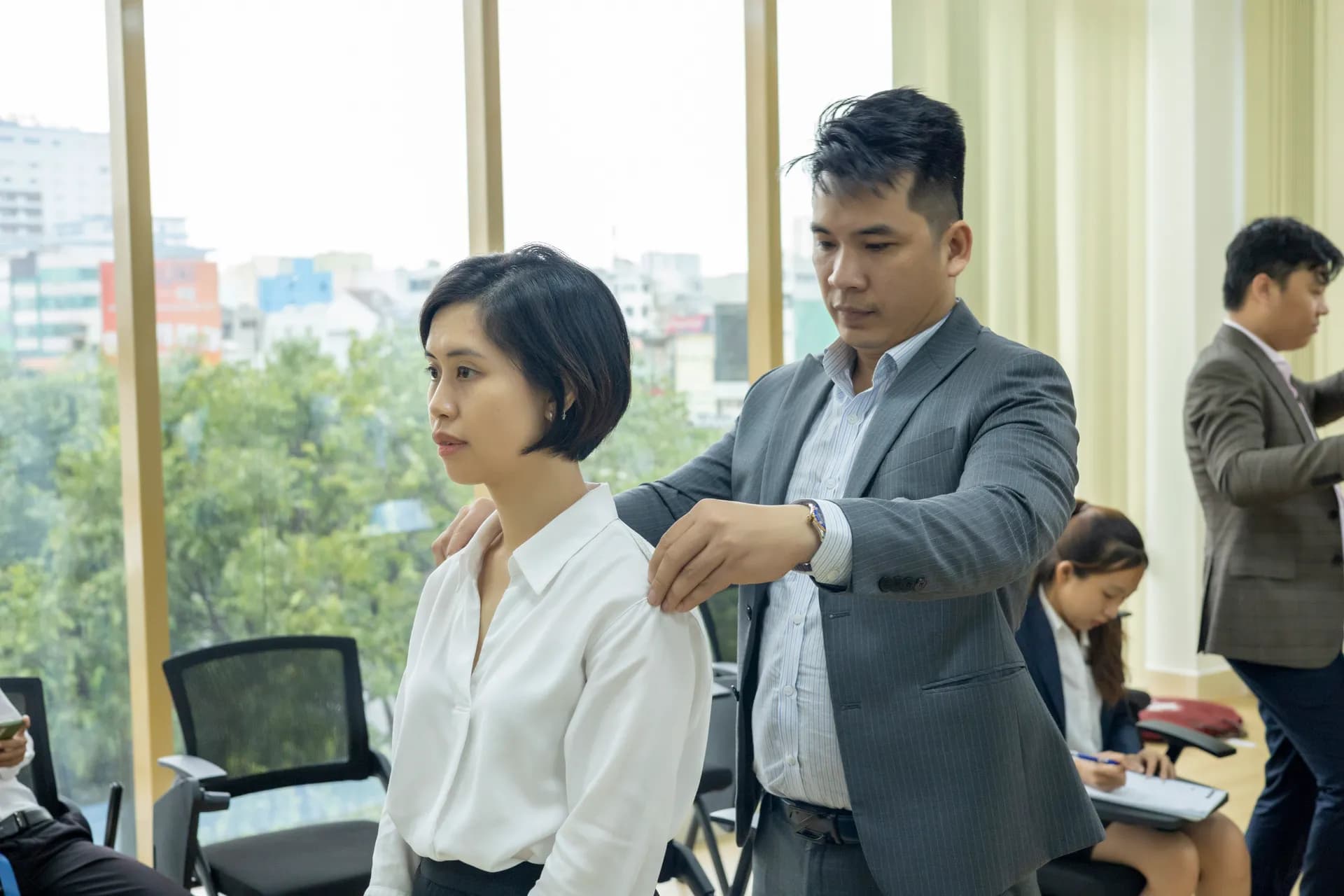 Thợ may vest xám đo vai khách nữ sơ mi trắng bên cửa sổ nhìn ra cây xanh
