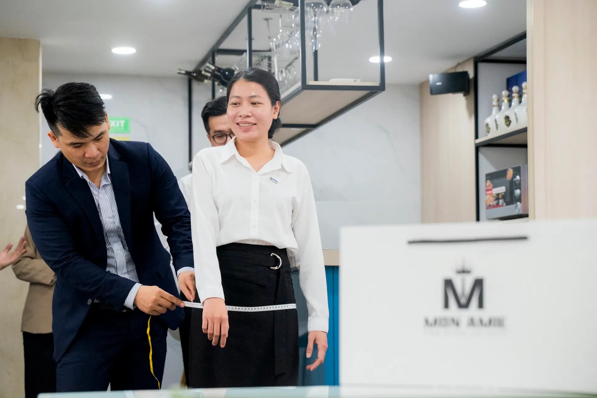 Khách nữ sơ mi trắng mỉm cười tự tin khi được đo vòng eo, túi giấy Mon Amie bên cạnh