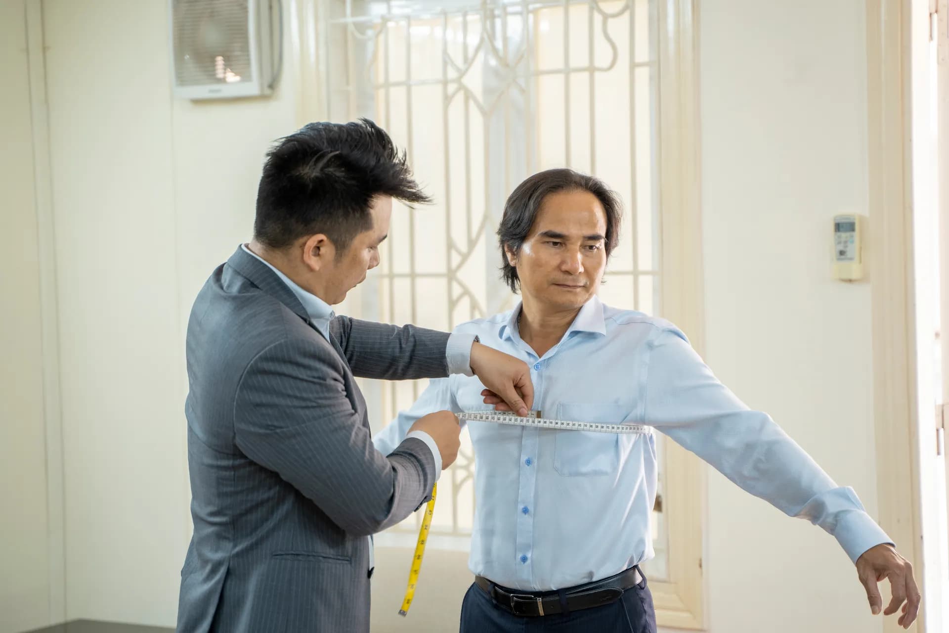Khách nam sơ mi xanh dang tay để thợ may vest xám đo vòng ngực, phòng sáng