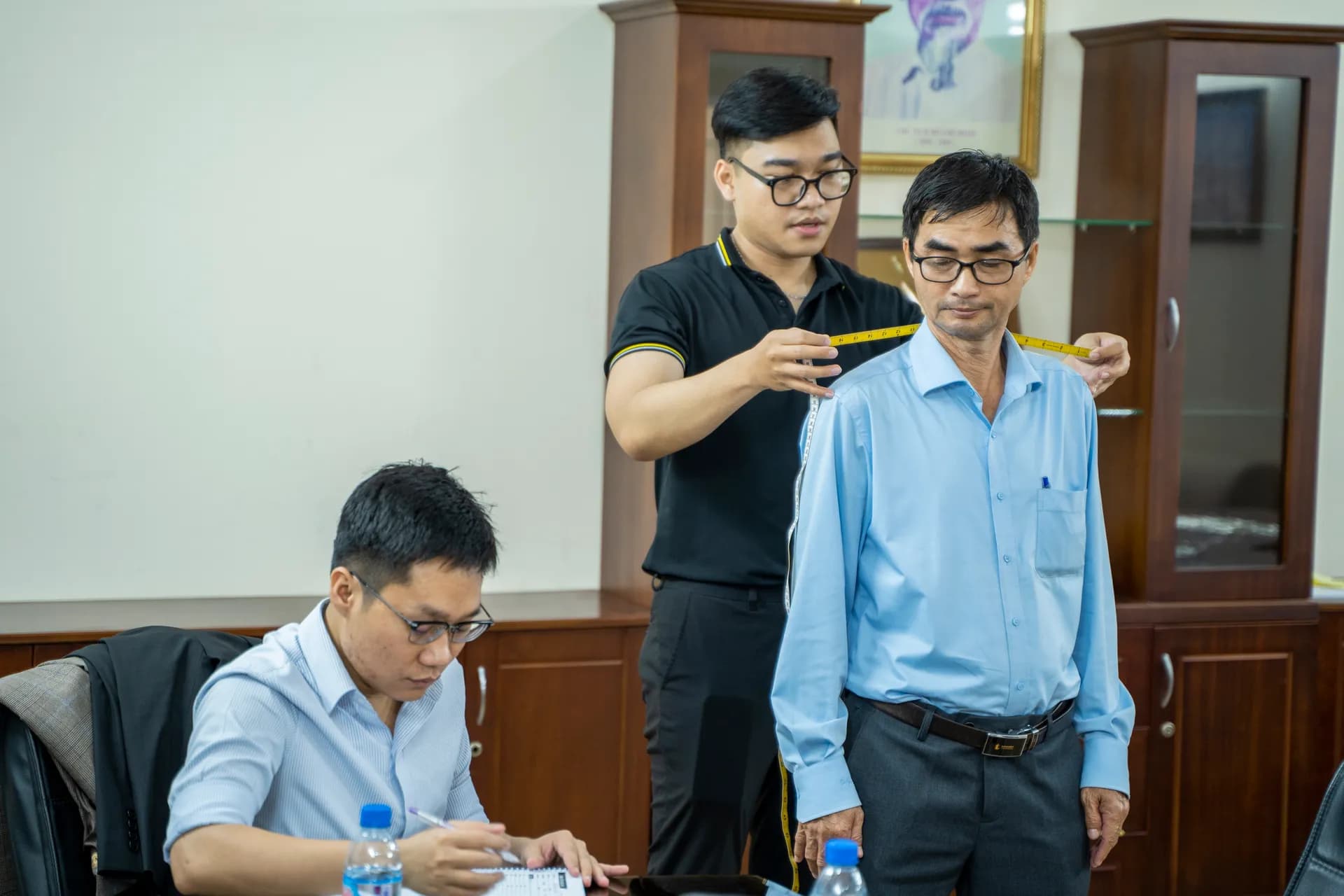 Thợ may áo polo đen đo vai cho khách nam sơ mi xanh nhạt trong phòng sáng