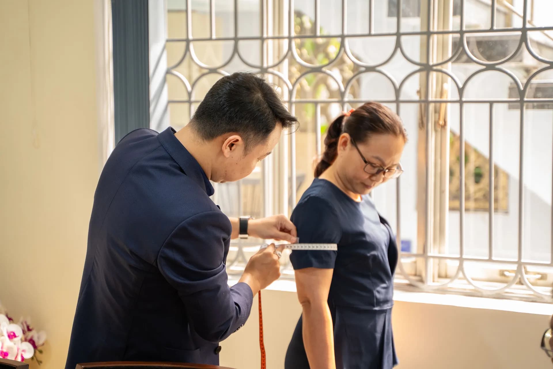 Thợ may vest navy đo bắp tay cho khách nữ váy xanh đậm bên cửa sổ