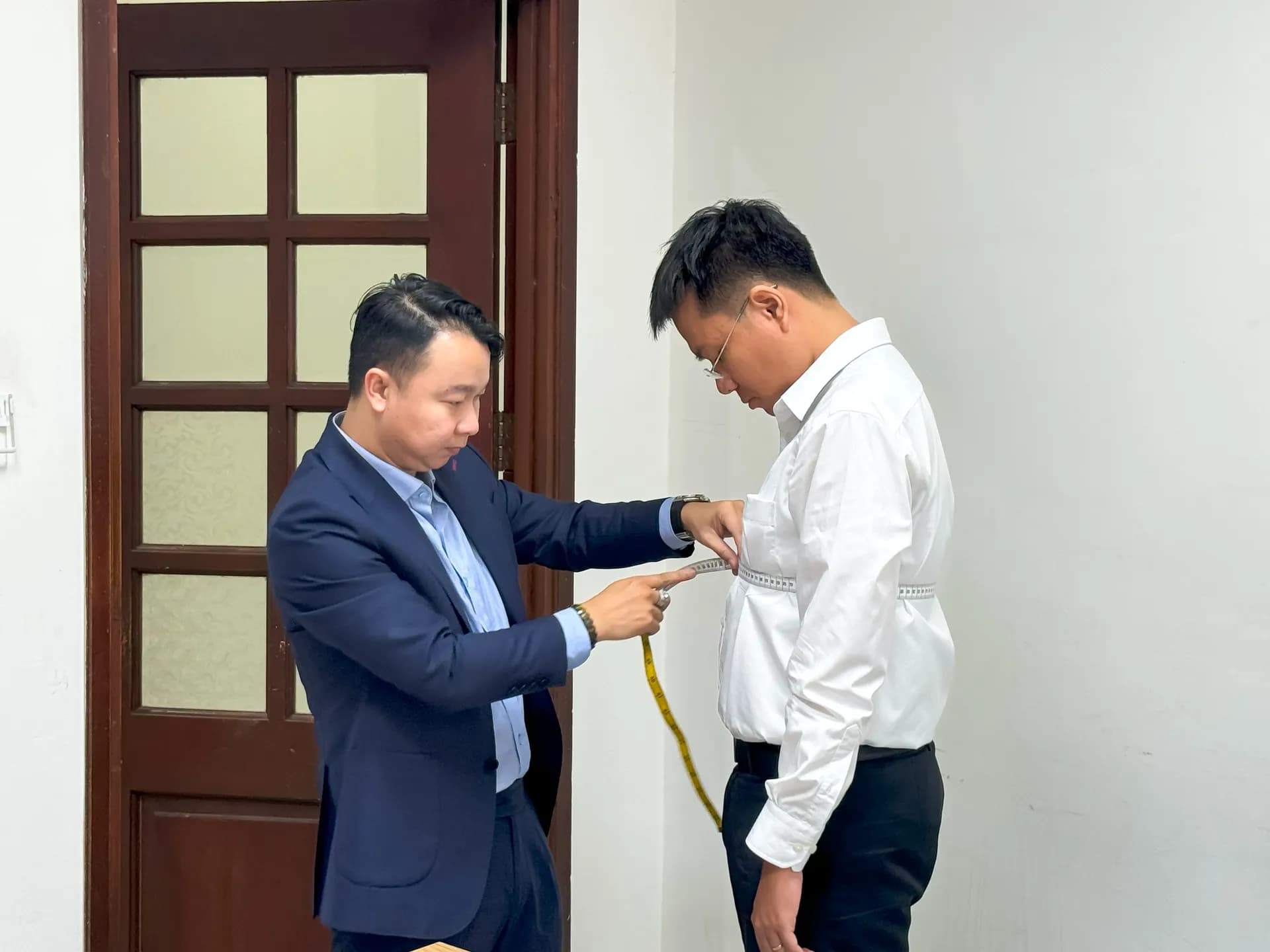 Thợ may vest navy đo vòng bụng cho khách nam sơ mi trắng bên cửa gỗ