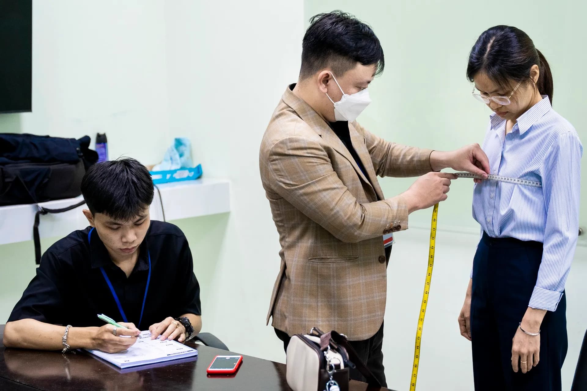 Đo vòng eo cho nữ nhân viên, trợ lý ngồi ghi số đo bên cạnh