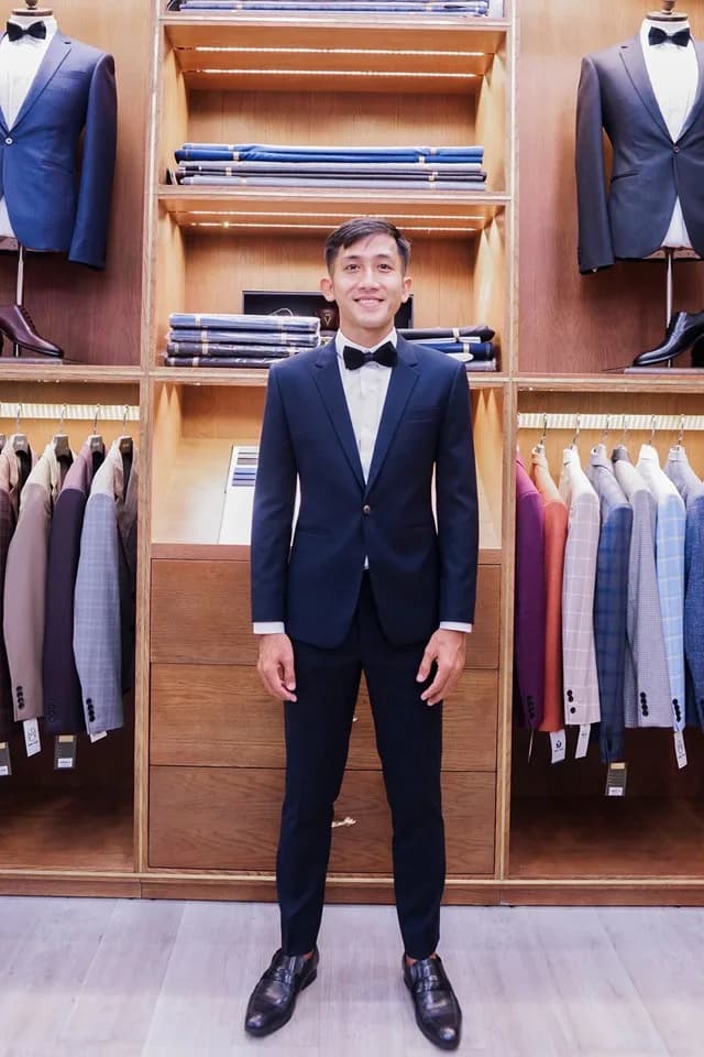 Suit xanh navy đậm với nơ đen, nụ cười rạng rỡ bên kệ vải gỗ: hình ảnh chú rể mà khách đi đám cưới nên tôn vinh, không lấn át