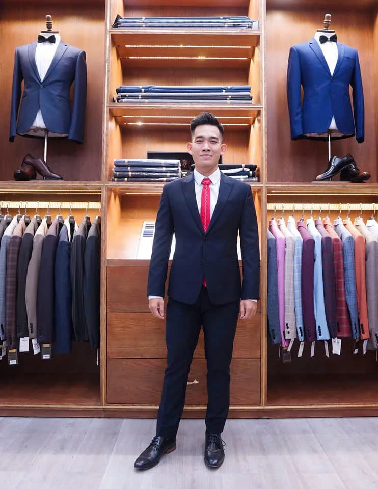 Chàng trai vest navy, cà vạt đỏ, đứng trong showroom gỗ sang trọng của tiệm may vest nam ở TP.HCM