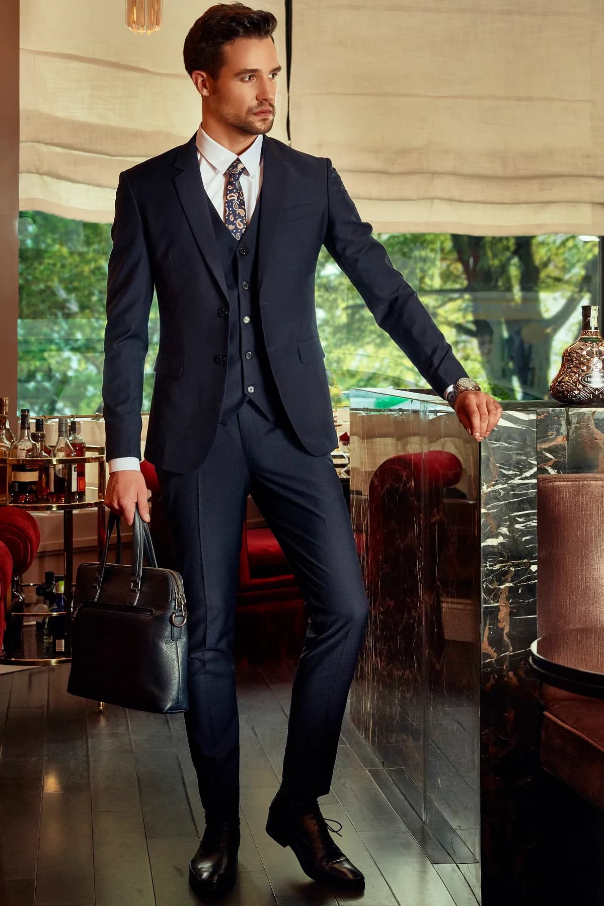 Doanh nhân trong suit navy ba mảnh, cà vạt paisley, tay xách cặp da, tựa quầy đá cẩm thạch, hình ảnh quà tặng doanh nhân cao cấp may đo