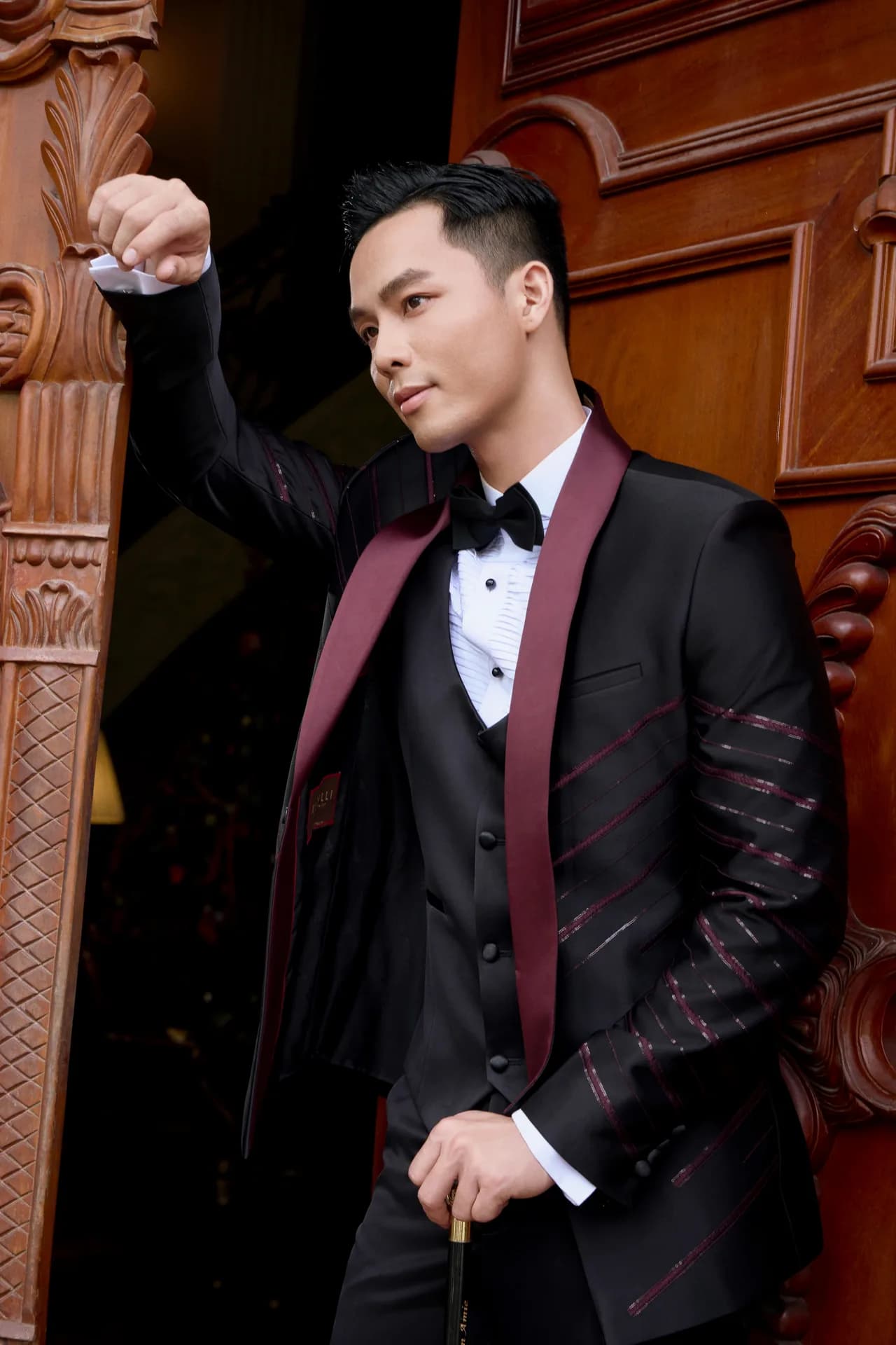 Tuxedo đen sọc kim tuyến đỏ rượu, ve shawl nhung đỏ, tựa cửa gỗ chạm khắc.