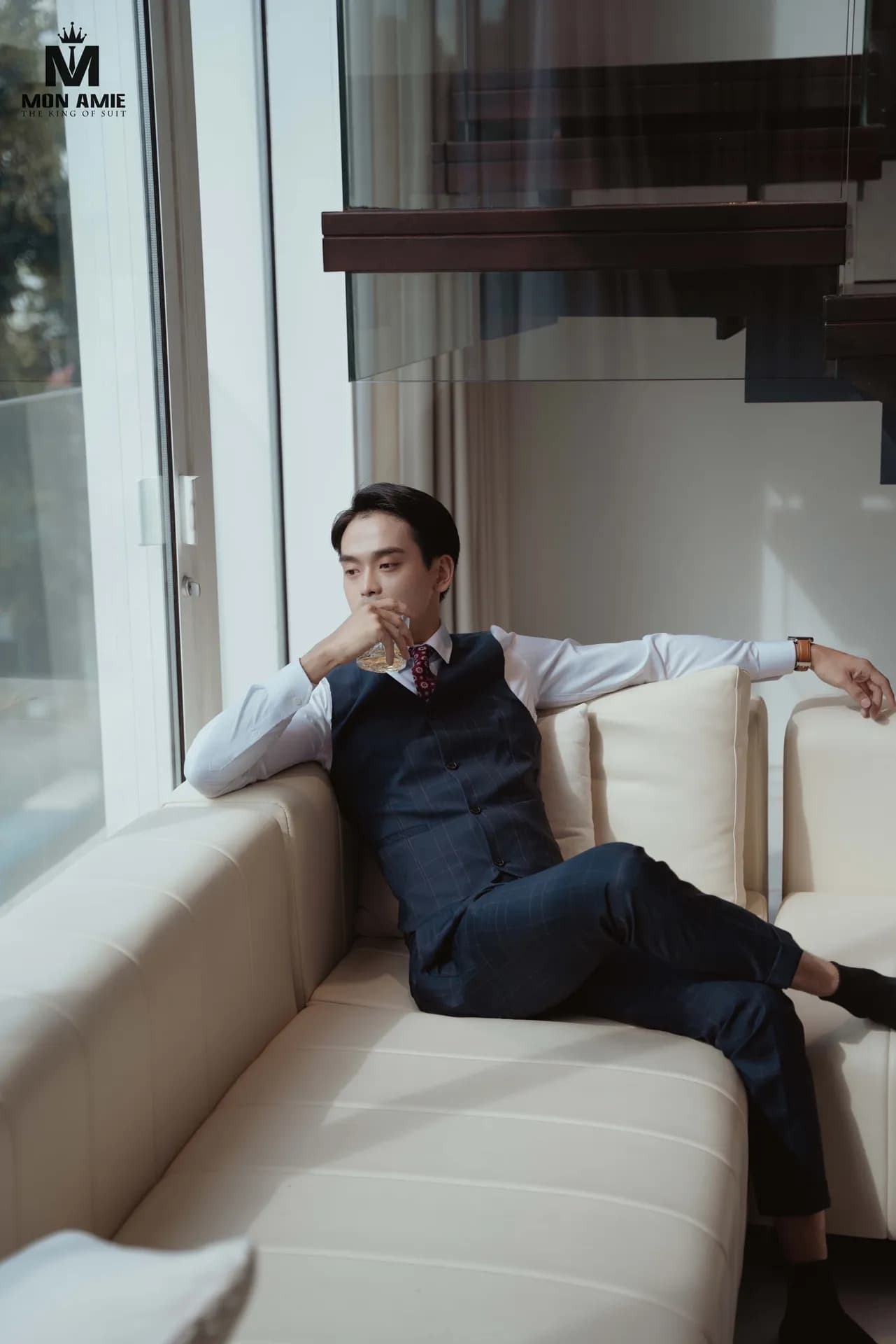 Gile caro xanh đen, sơ mi trắng, thư giãn trên sofa kem cạnh cửa kính