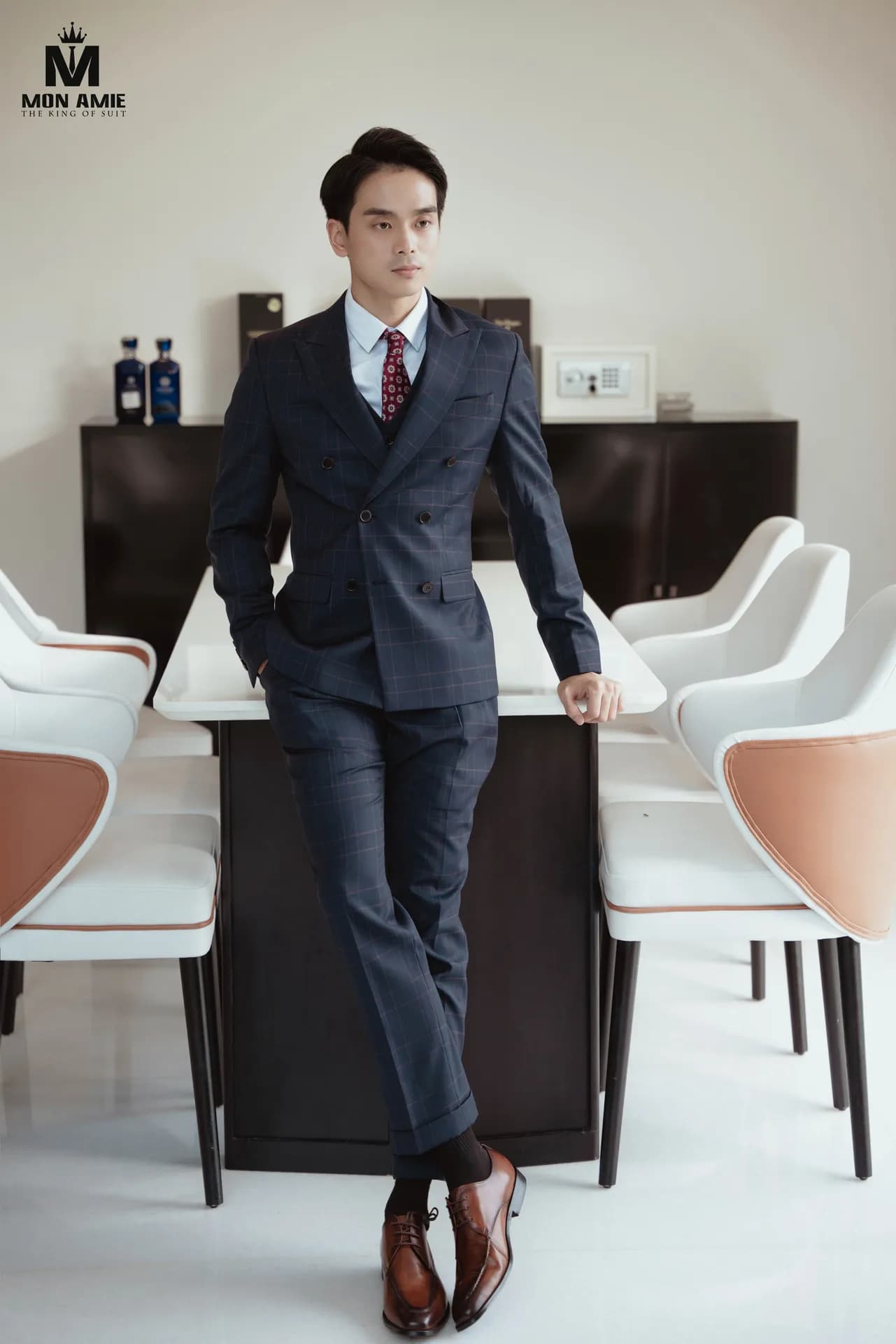 Suit hai hàng khuy caro xanh đen ba mảnh, dựa bàn họp trắng