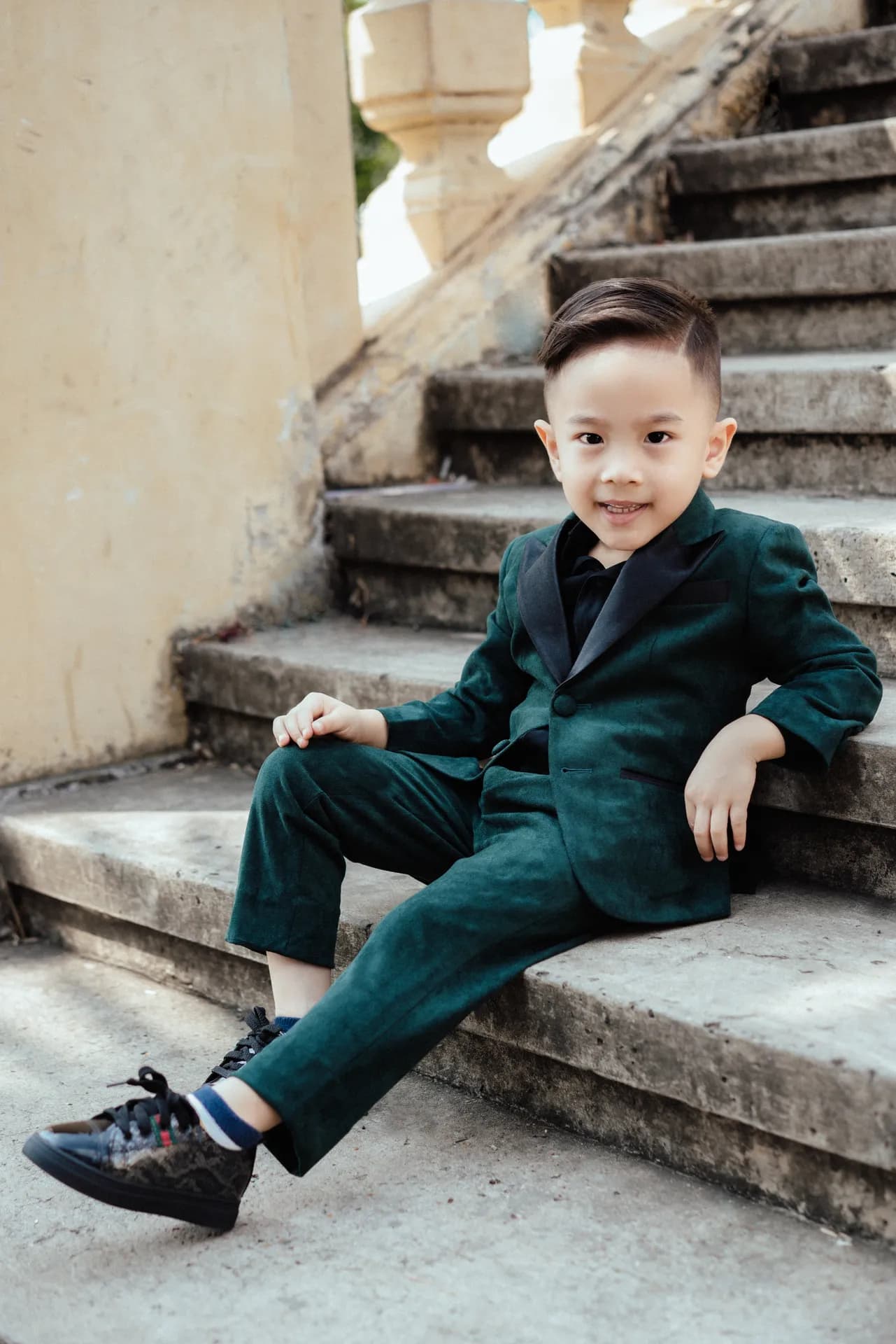 Bé trai mặc suit nhung xanh lục cổ lụa đen ngồi trên bậc thang
