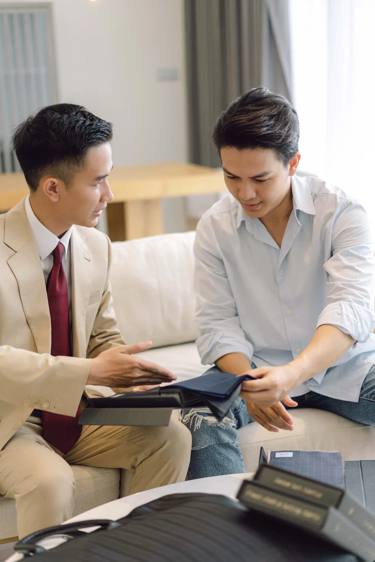 Thợ may vest be, cà vạt đỏ rượu, giới thiệu sổ mẫu vải cho khách tại phòng khách