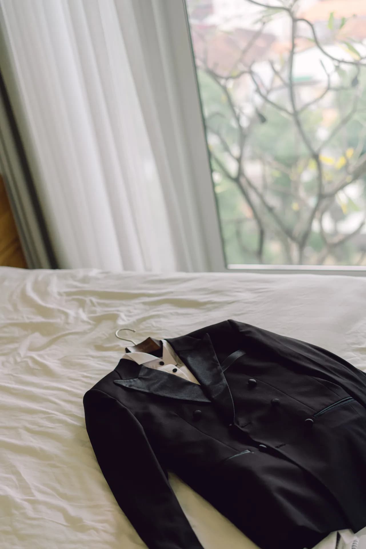 Bộ tuxedo đen và sơ mi trắng đặt trên giường cạnh cửa sổ
