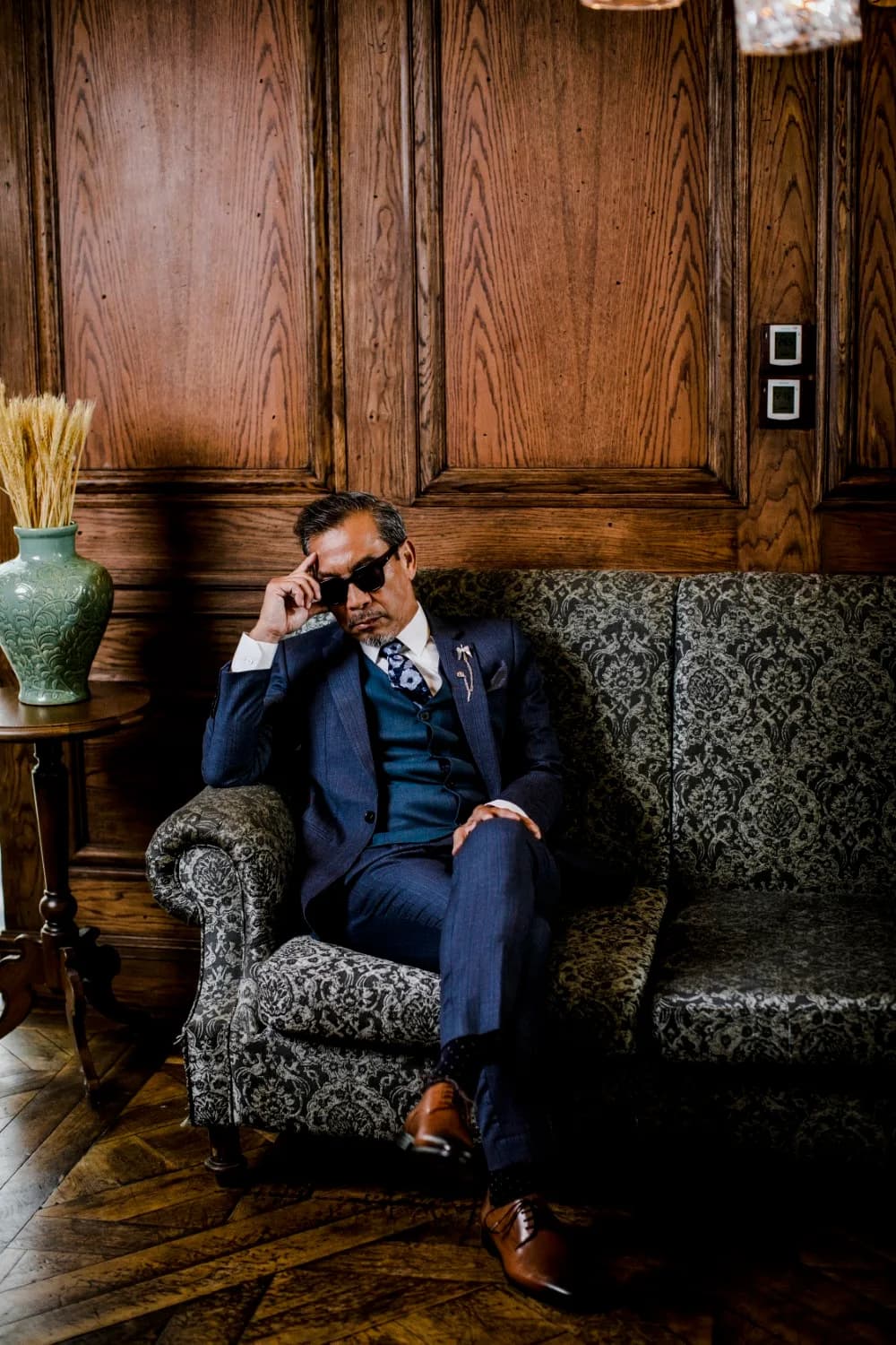 Suit navy kẻ sọc mảnh, gilê xanh ngọc, ngồi ghế bọc họa tiết, ví dụ vest cho người béo với sọc dọc mảnh và form gọn