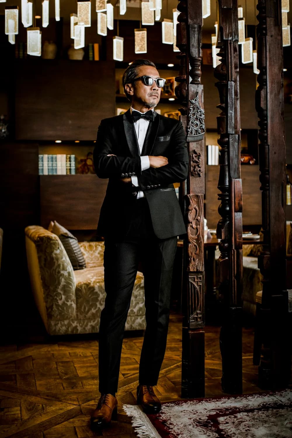 Tuxedo đen toàn thân, nơ đen, cài áo, đứng giữa cột gỗ chạm