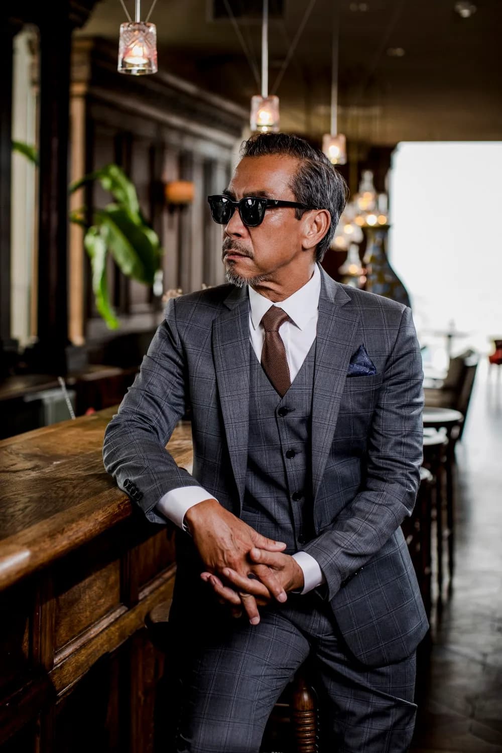 Suit ba mảnh kẻ ô xám xanh, cà vạt nâu, chắp tay ngồi tựa quầy bar