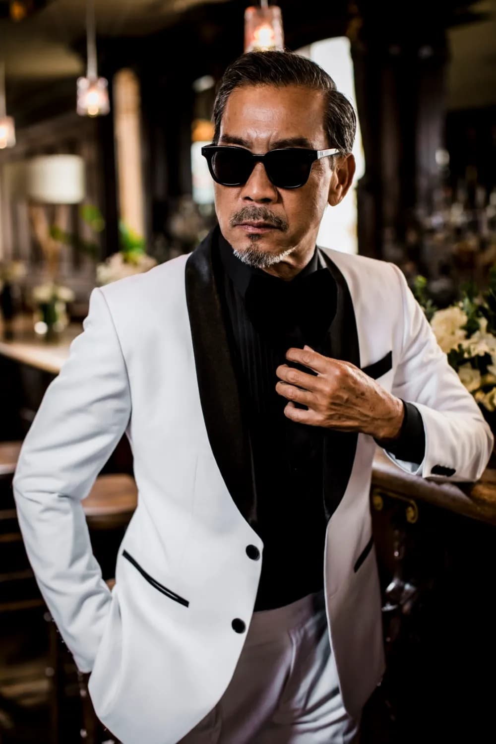 Tuxedo trắng ve nhung đen, kính râm, tay đút túi bên quầy bar
