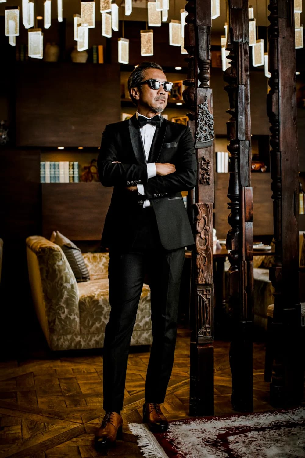 Tuxedo nhung đen thắt nơ, khoanh tay trong không gian gỗ chạm ánh đèn