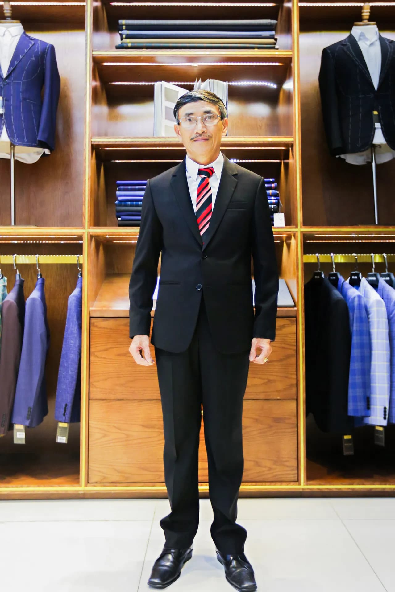 Suit đen hai cúc, cà vạt sọc đỏ đen nổi bật, đứng thẳng trước kệ gỗ