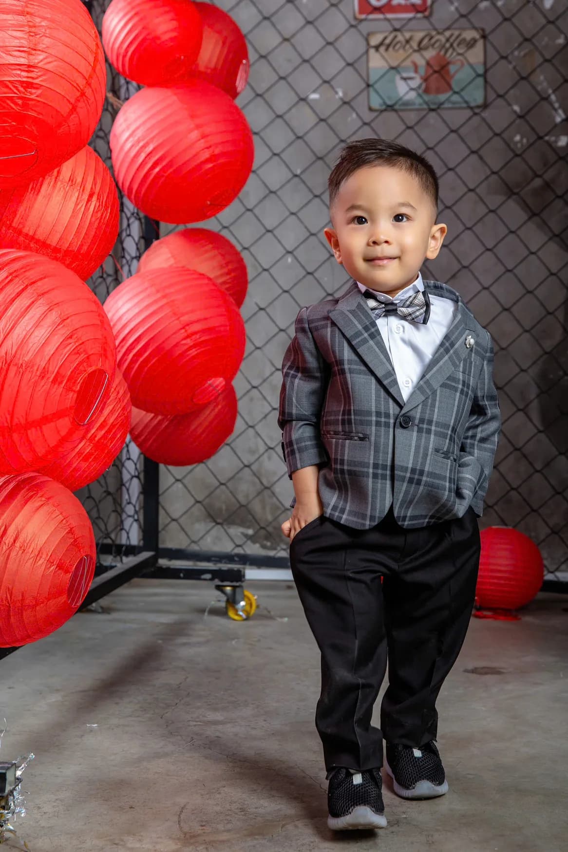 Bé trai vest caro xám, nơ, quần đen, nền lồng đèn đỏ