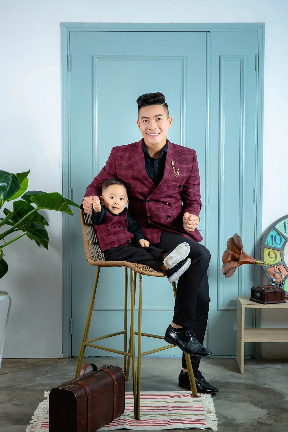 Bố con vest caro đỏ mận, ngồi ghế mây, nền cửa xanh