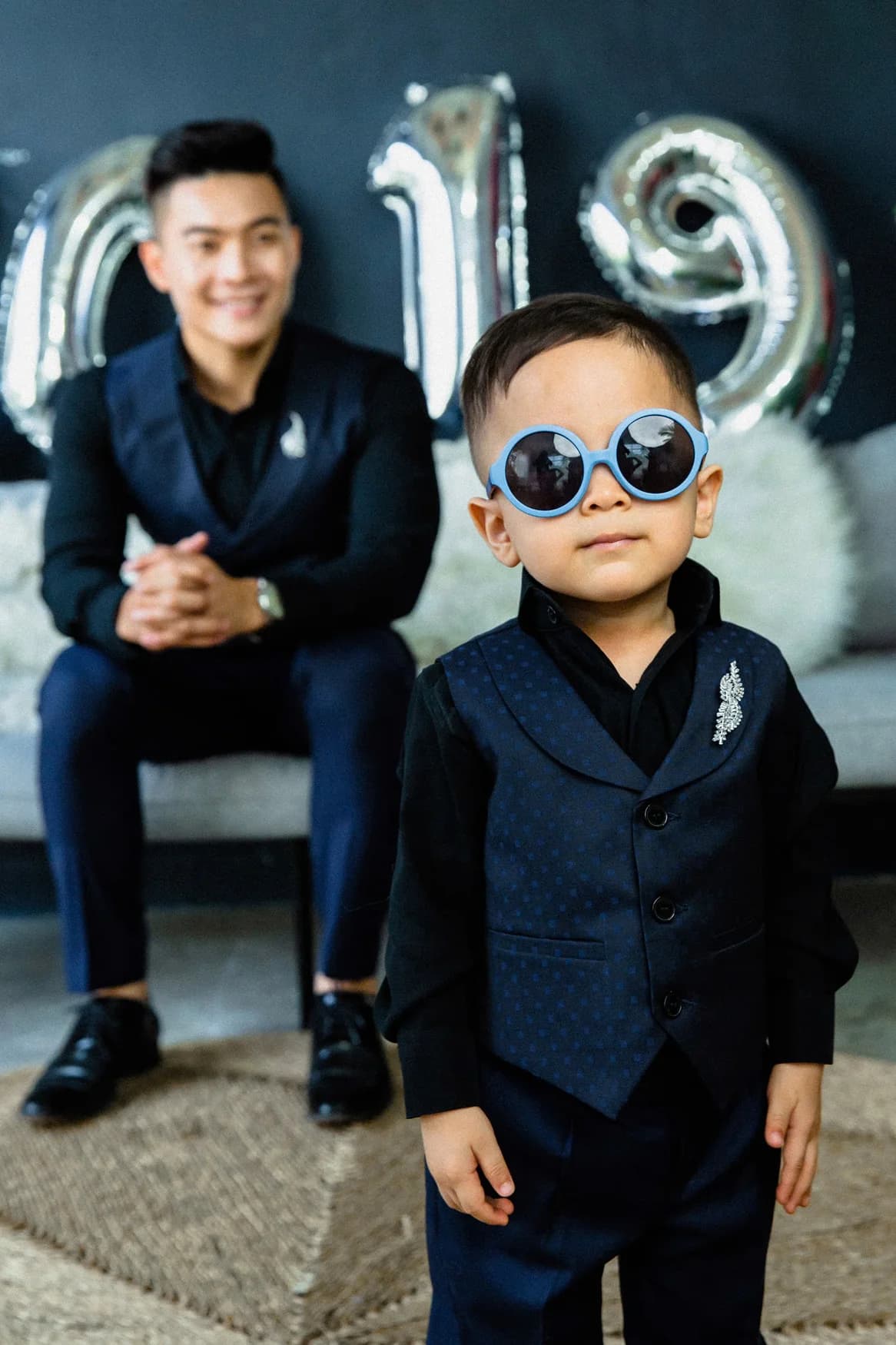 Bé trai gile xanh navy, kính mát xanh, bố ngồi phía sau