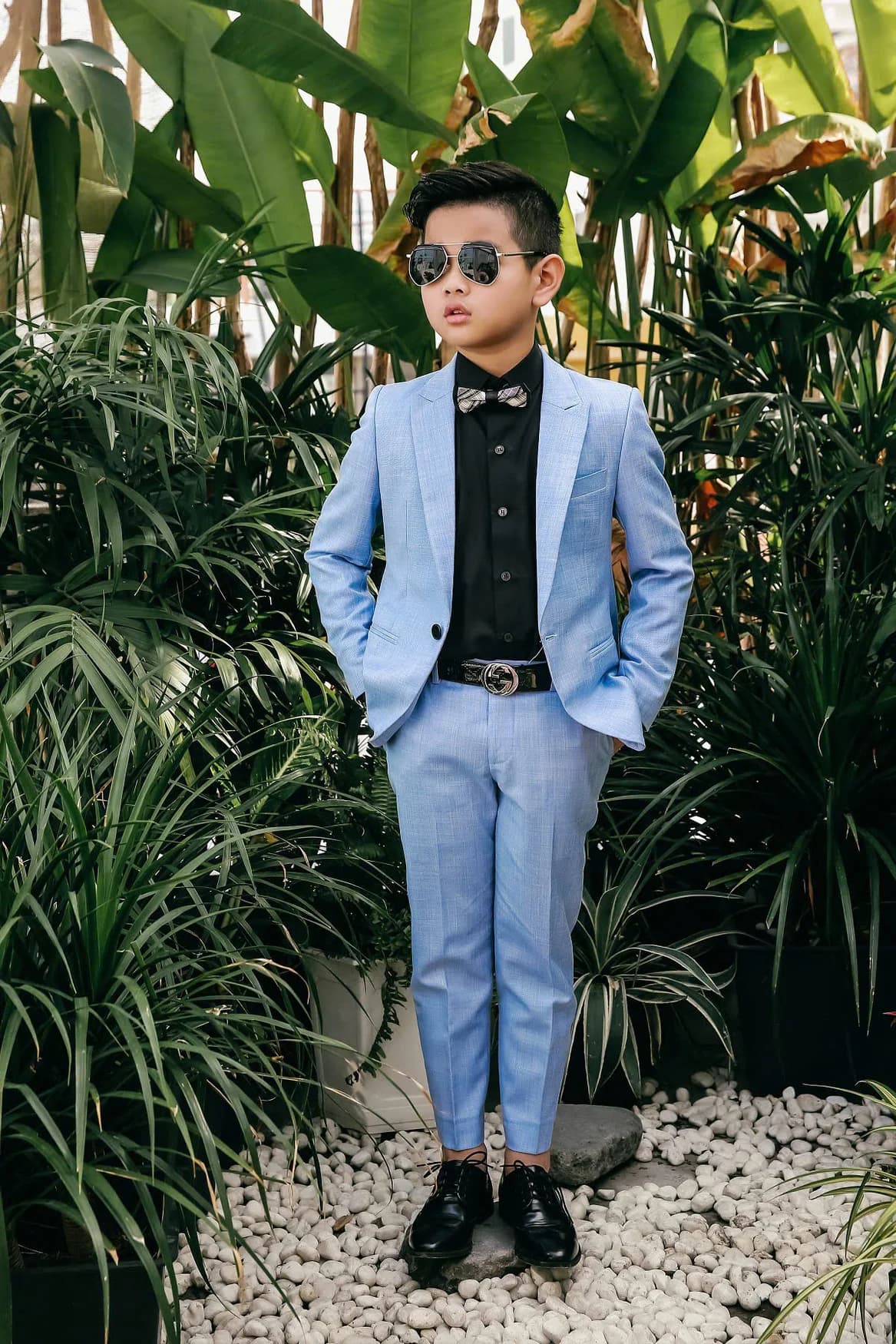 Bé trai vest xanh pastel, sơ mi đen, kính râm, sân vườn