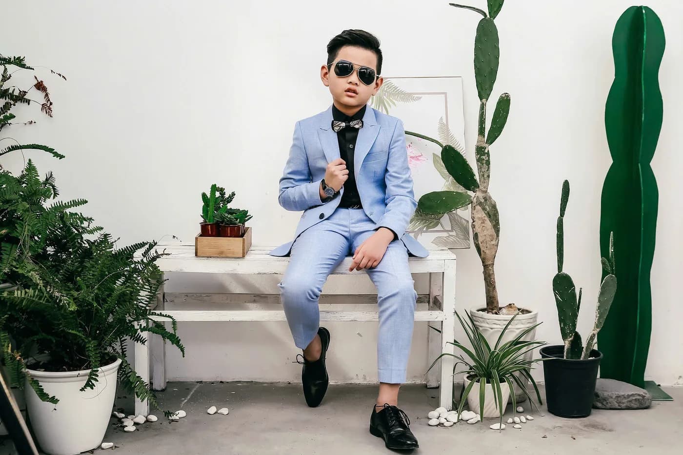 Bé trai vest xanh pastel ngồi ghế trắng, kính râm, xương rồng