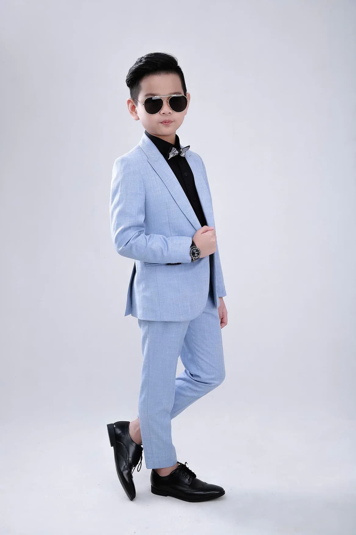 Bé trai vest xanh pastel nghiêng người, kính râm, nền trắng