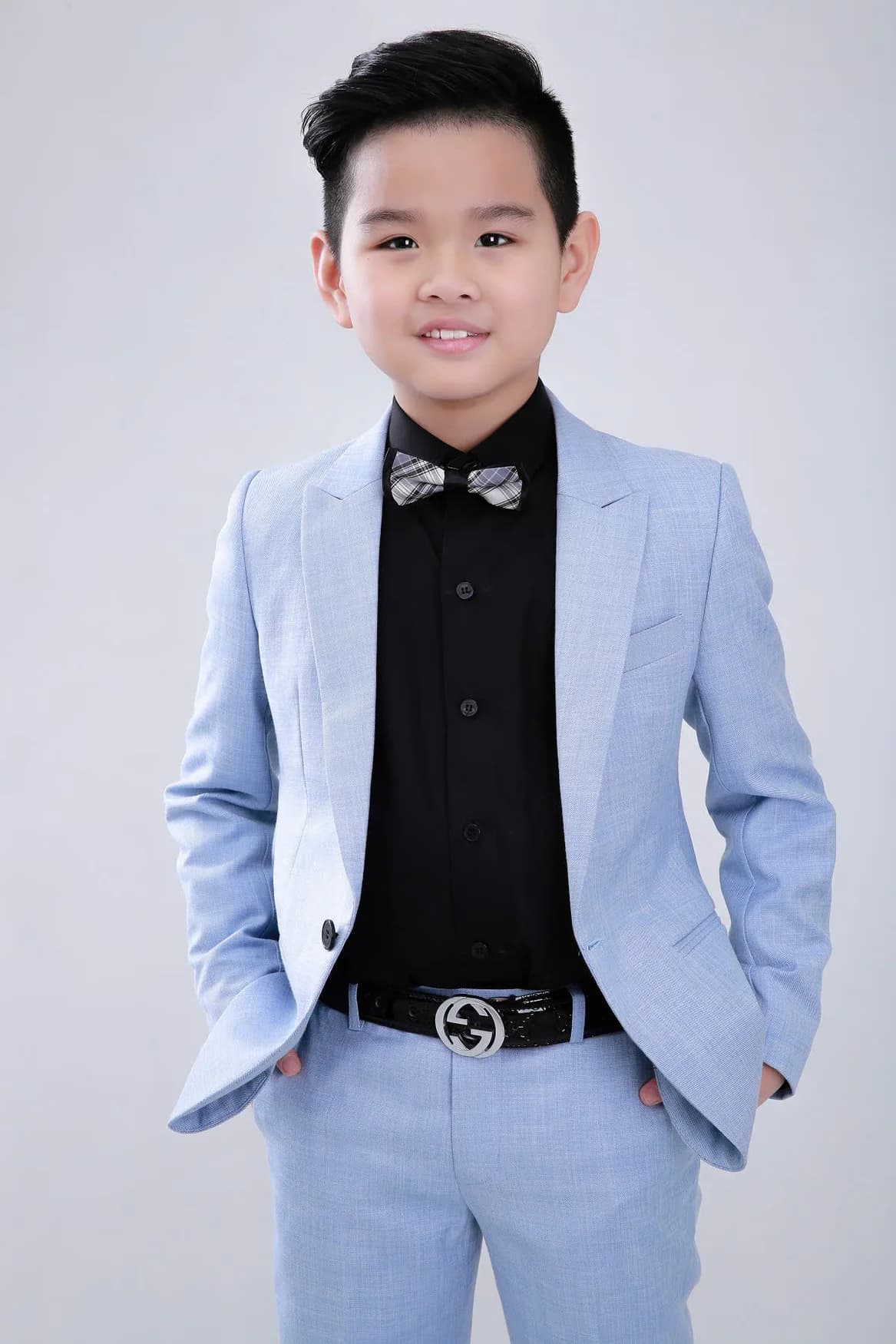 Bé trai cười, vest xanh pastel, sơ mi đen, nơ caro