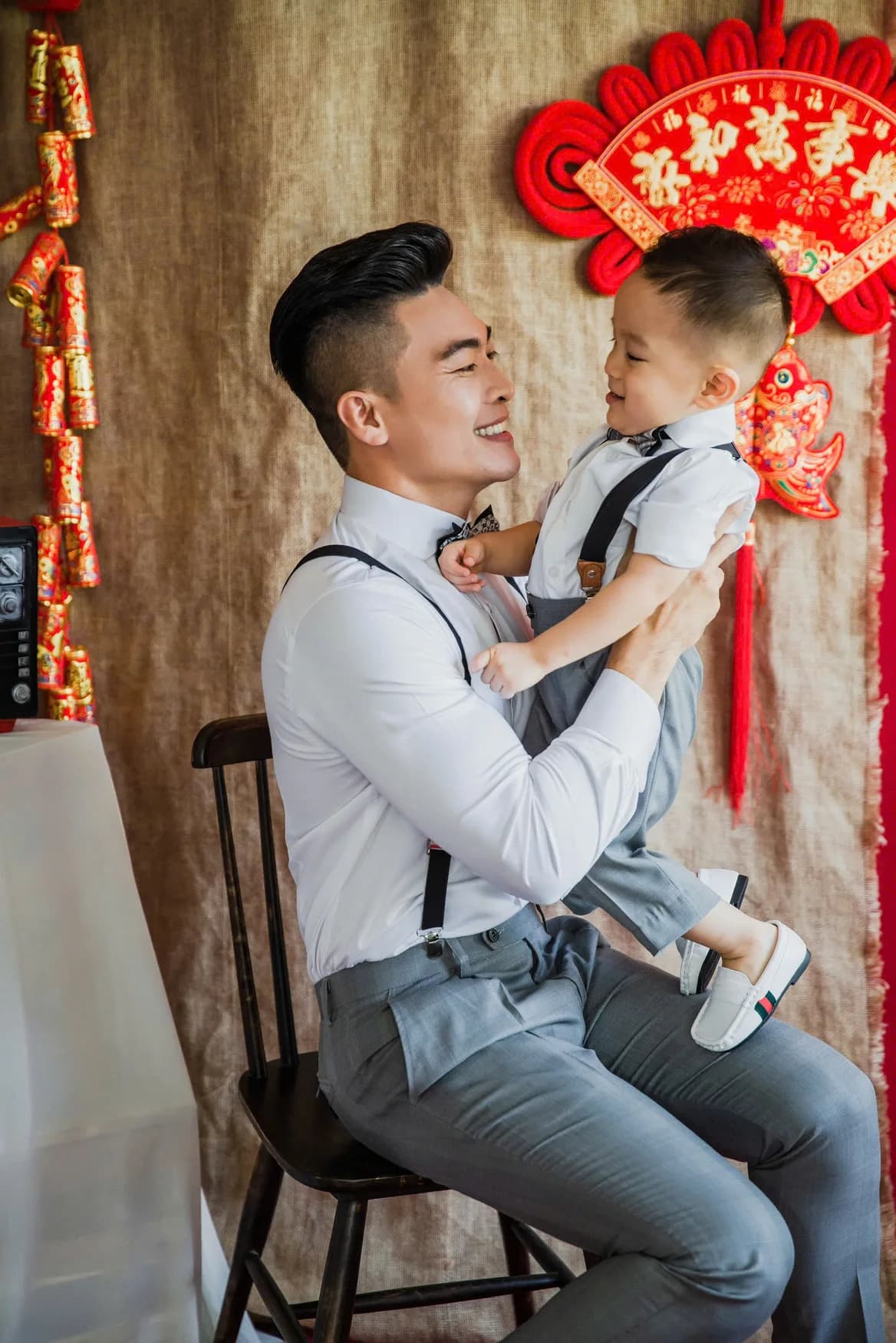 Bố sơ mi trắng bế bé quần yếm xám, phông Tết đỏ