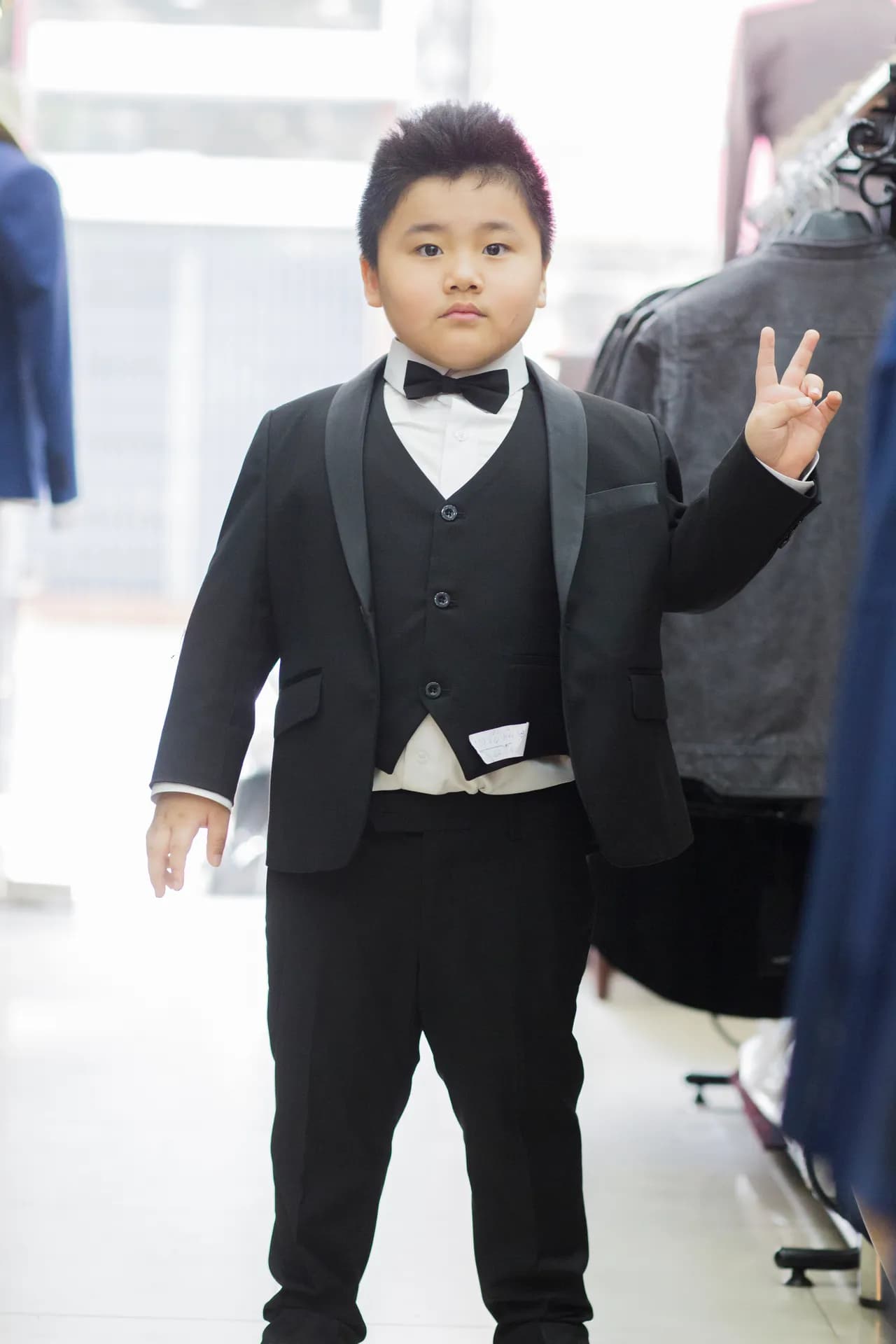 Bé trai mặc tuxedo đen ba mảnh, giơ tay chữ V dễ thương