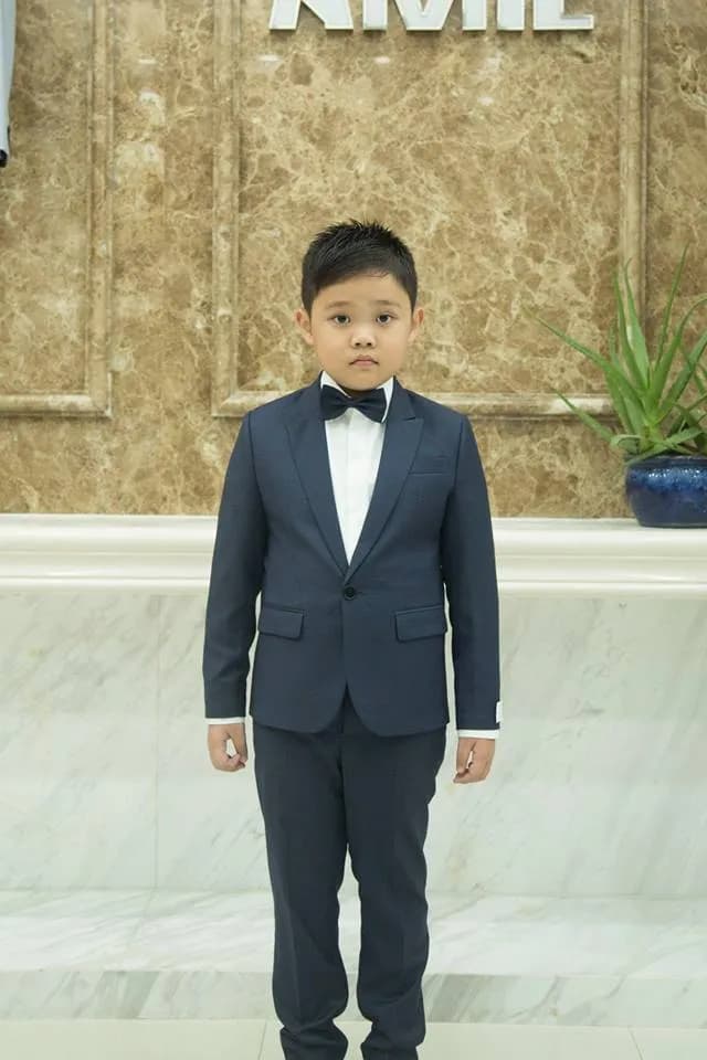 Suit xanh navy, nơ đen, dáng đứng nghiêm chỉnh trước tường đá cẩm thạc