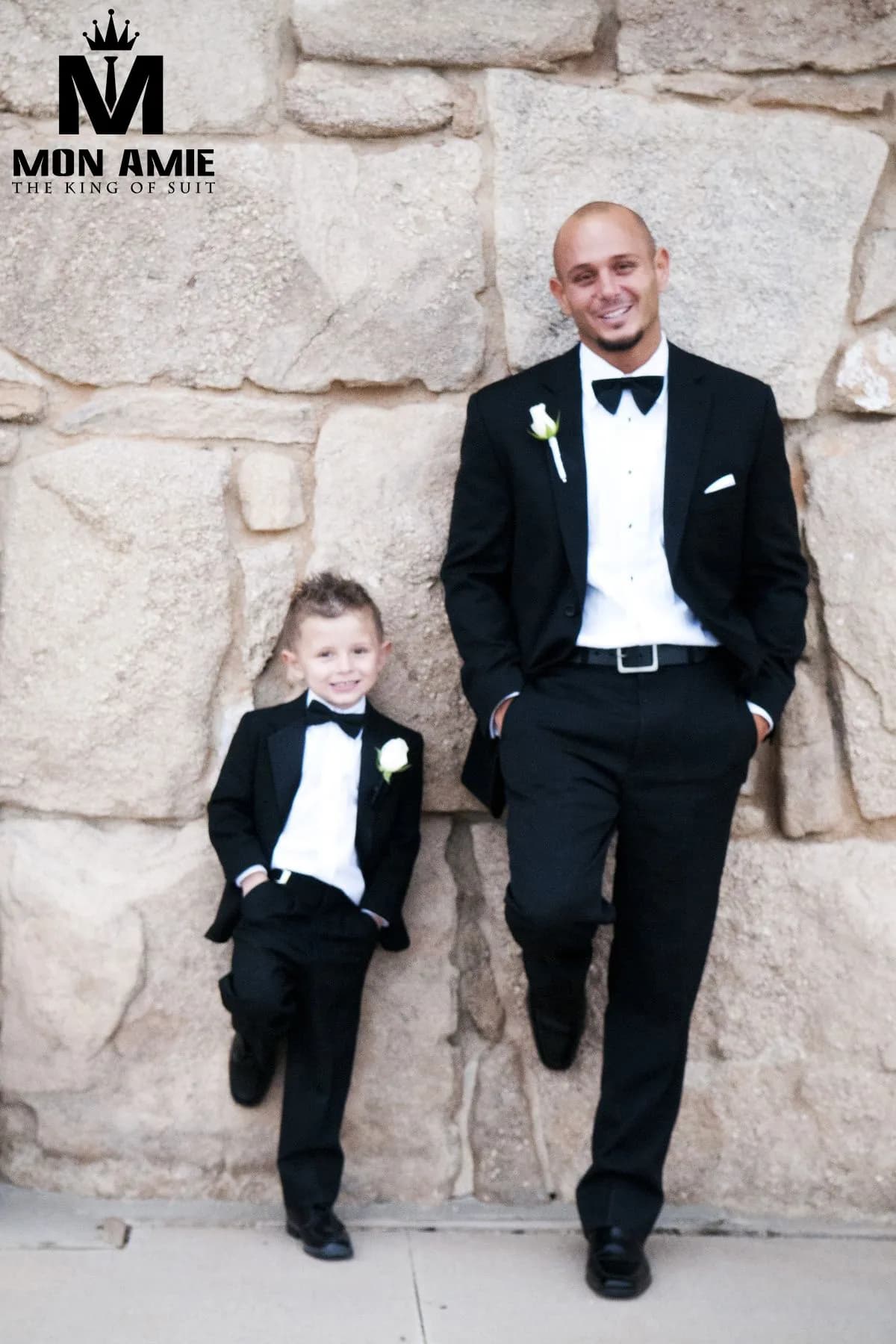 Người lớn và cậu bé cùng tuxedo đen, dựa tường đá với nụ cười tươi
