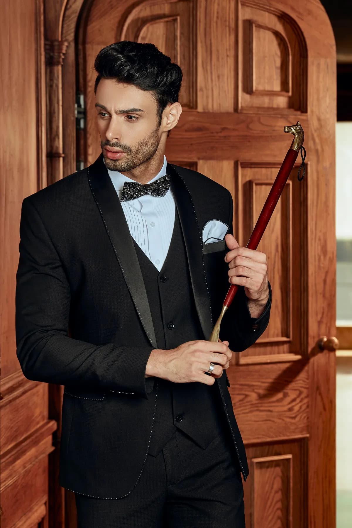 Tuxedo đen ba mảnh ve satin, nơ, tay cầm gậy đỏ bên cửa gỗ vòm