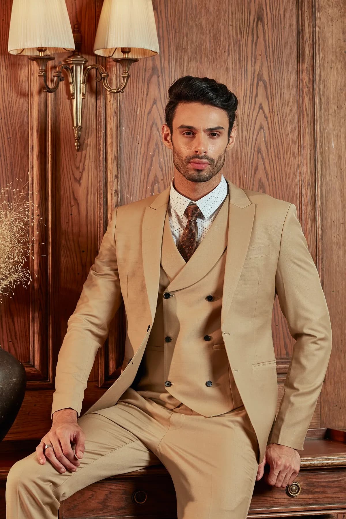 Suit ba mảnh be camel, gilê hai hàng khuy, ngồi tựa bàn gỗ