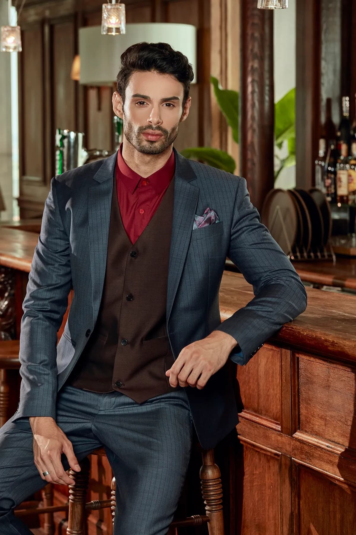 Suit xanh khói, gilê nâu, sơ mi đỏ rượu, ngồi ghế cao bên quầy bar