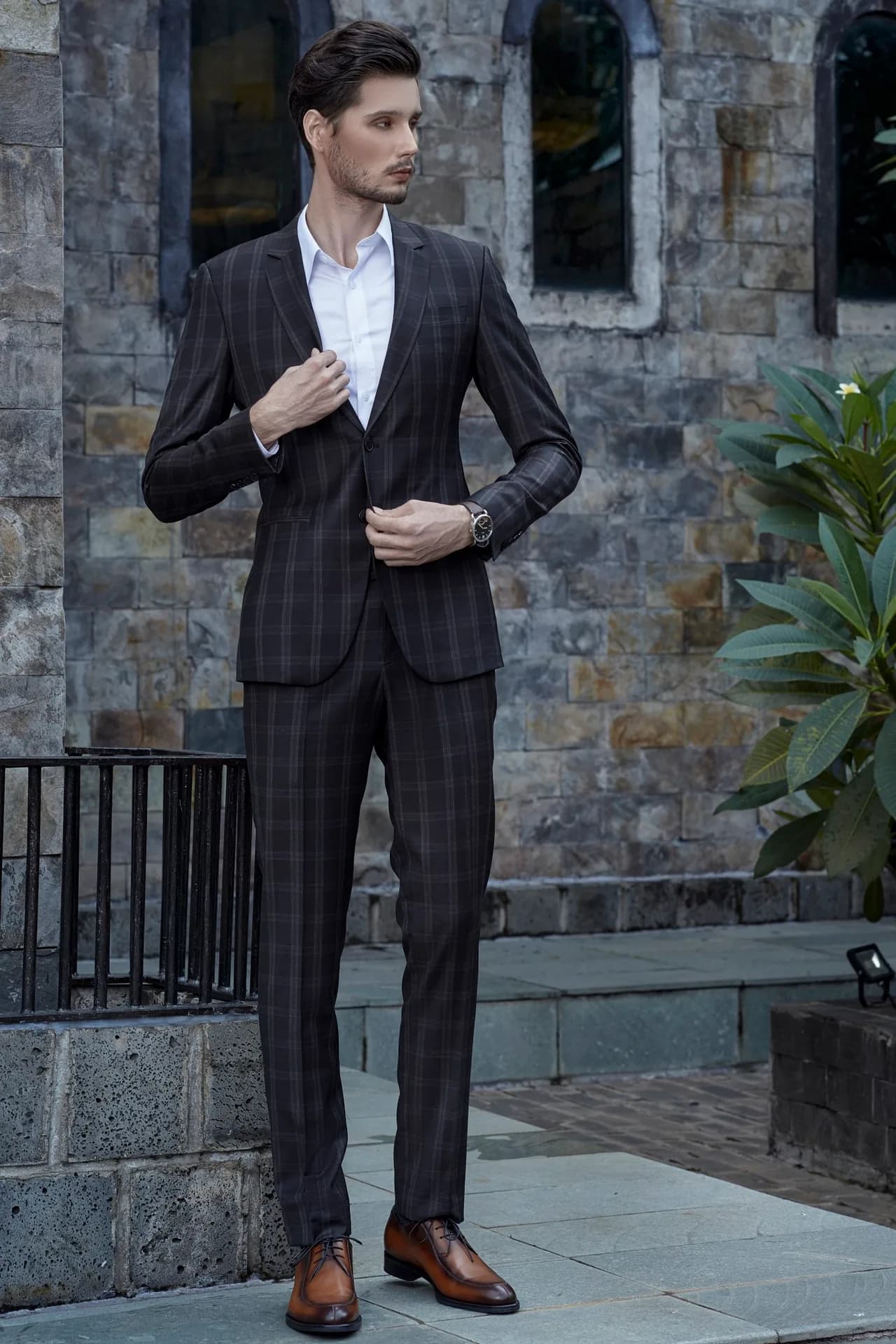 Suit caro nâu đen dáng đứng trước tường đá, sơ mi trắng cổ đứng, minh họa trang phục lịch sự cho nam