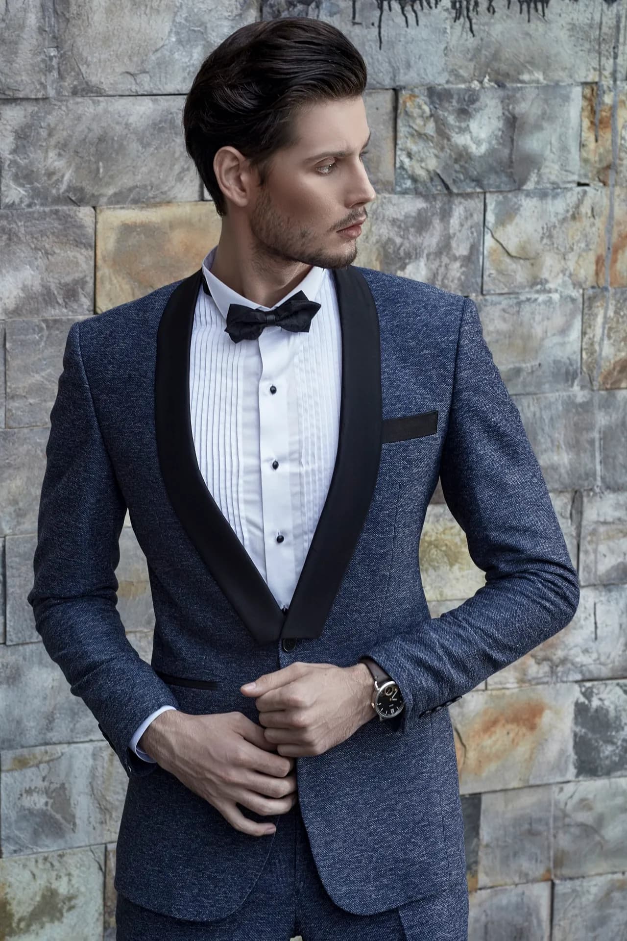 Tuxedo xanh navy ve nhung đen, nơ, sơ mi xếp ly, cận cảnh trước tường đá