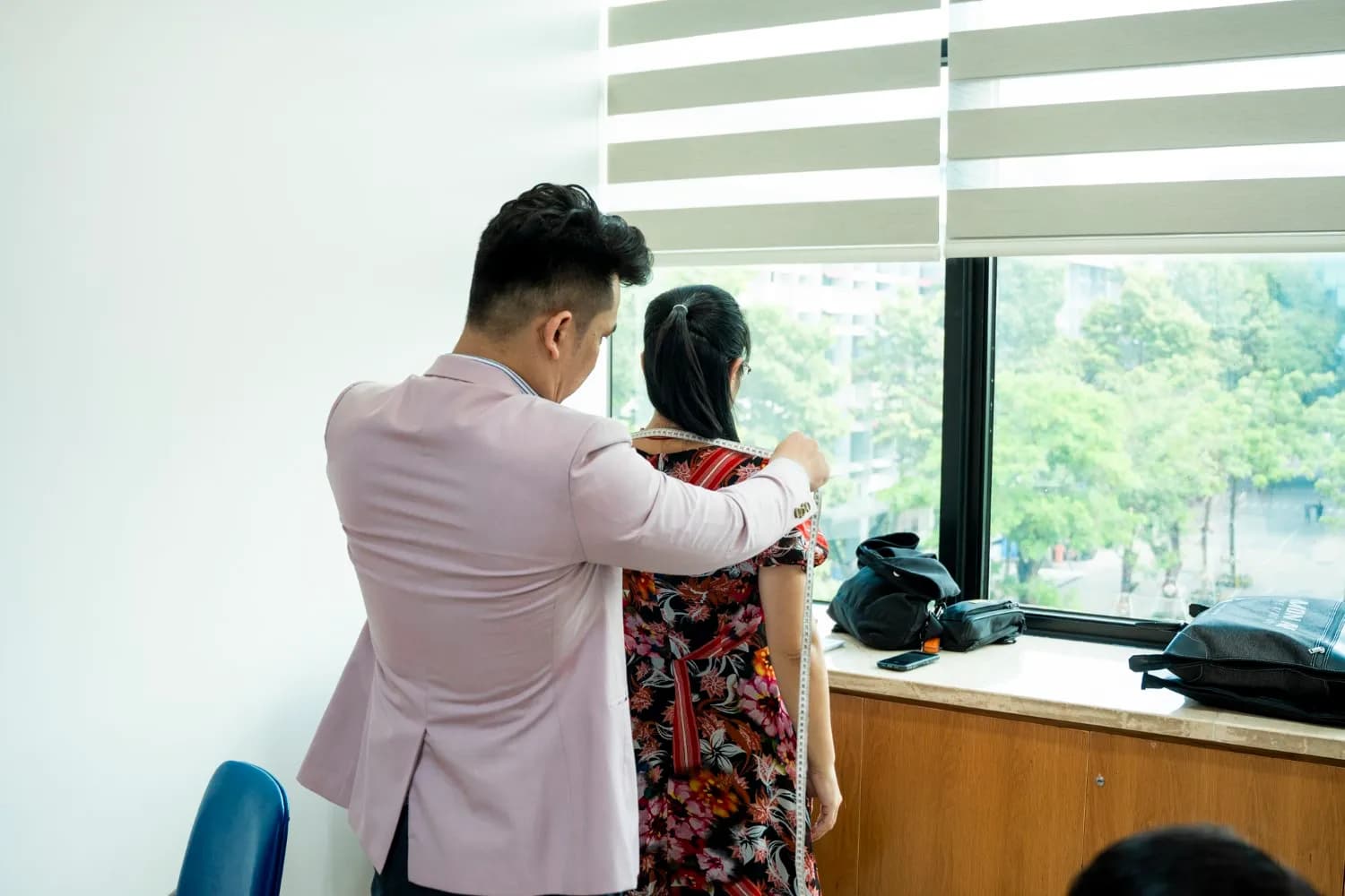 Thợ blazer hồng đo vai cho khách nữ váy hoa bên cửa sổ sáng nhìn ra cây xanh