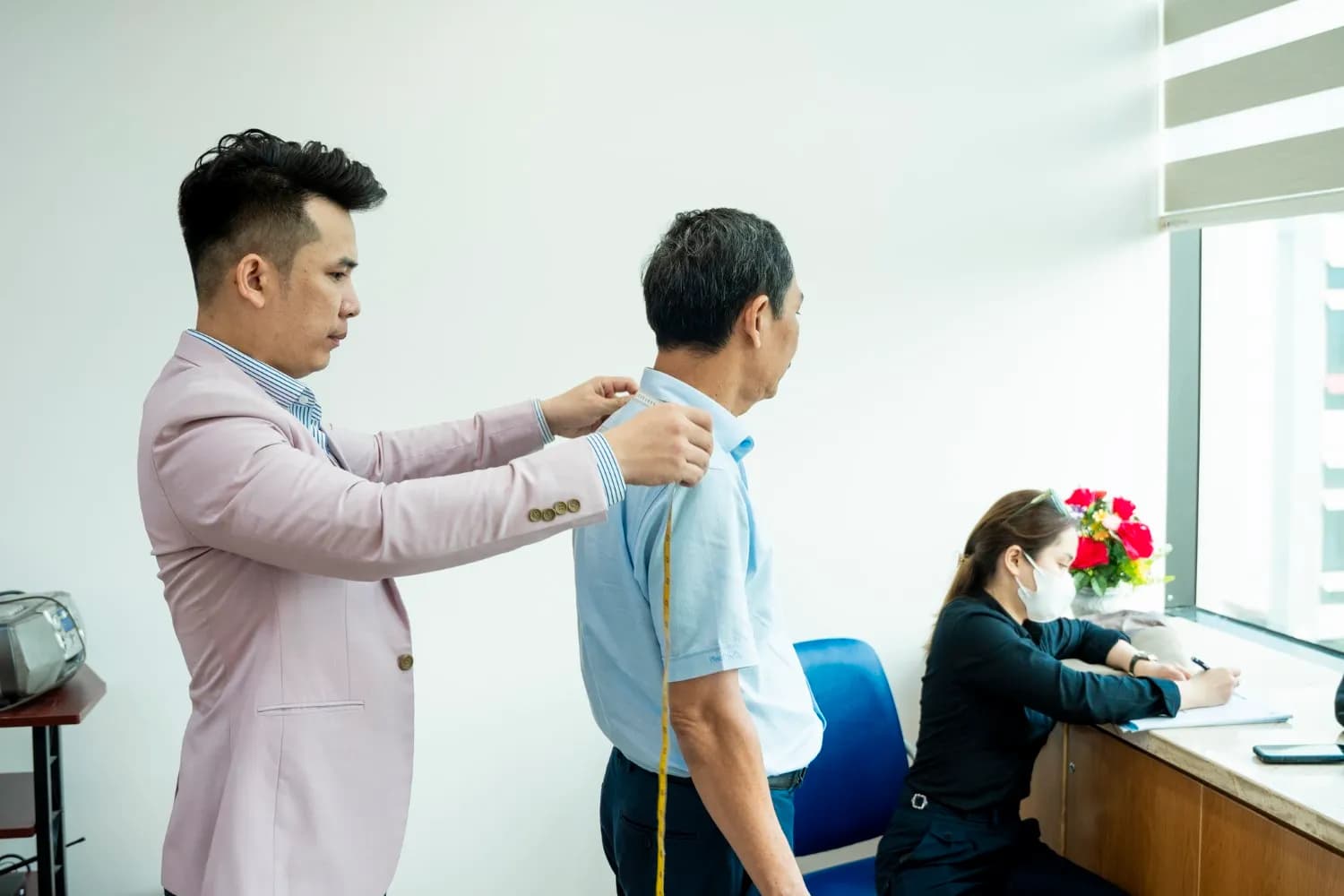 Đo chiều dài tay áo cho khách sơ mi xanh nhạt, trợ lý ghi số đo bên bàn hoa hồng đỏ