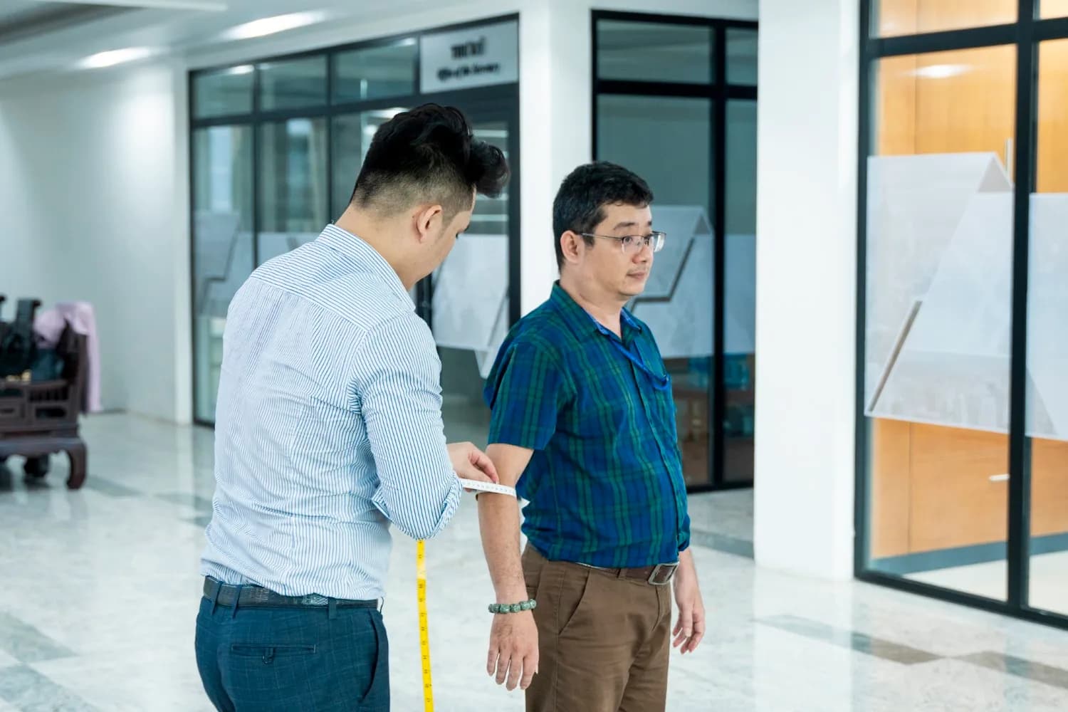 Đo tay áo cho khách sơ mi kẻ xanh trong sảnh sáng rộng, thước dây vàng