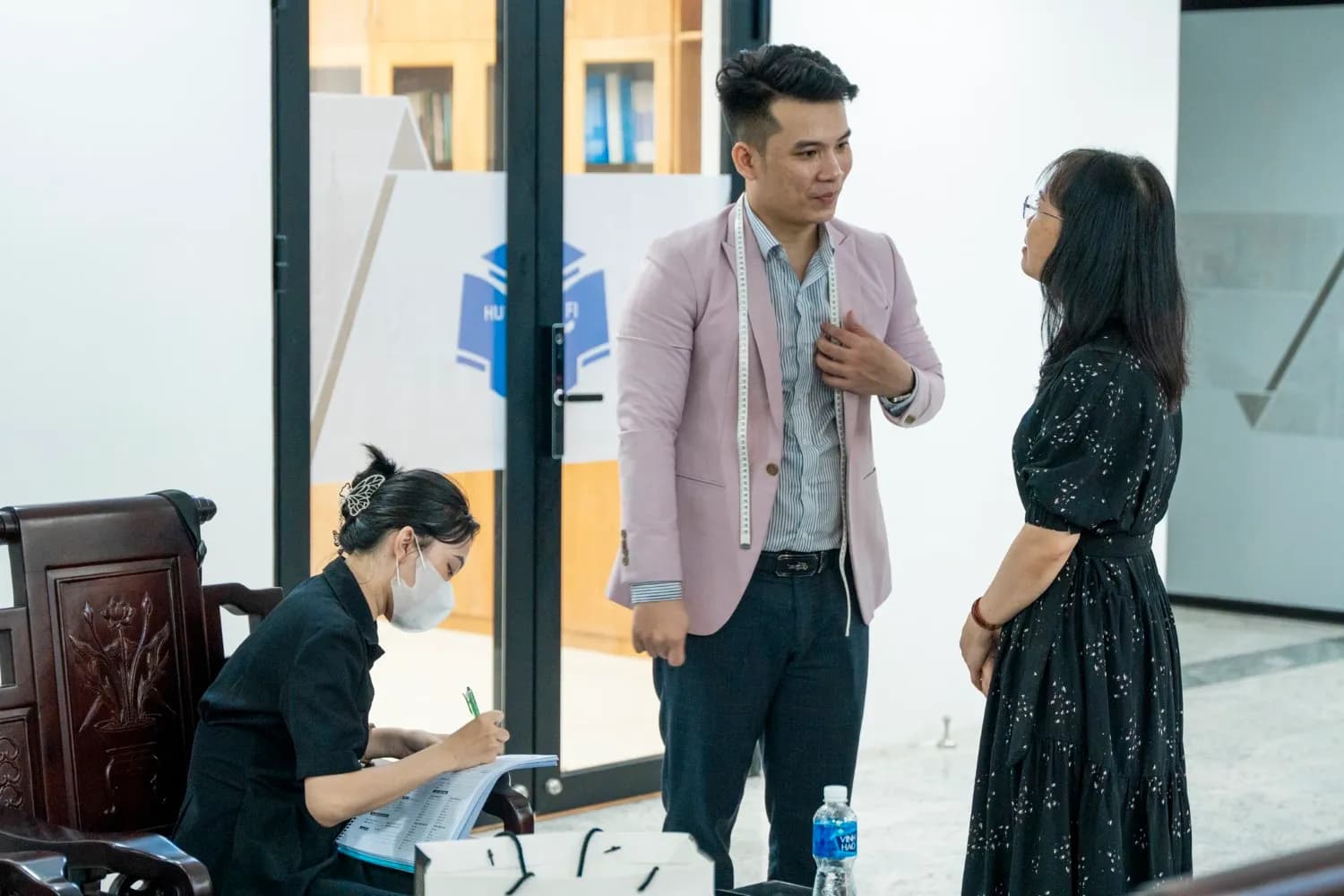 Thợ blazer hồng vắt thước dây quanh cổ, trao đổi với khách nữ váy đen