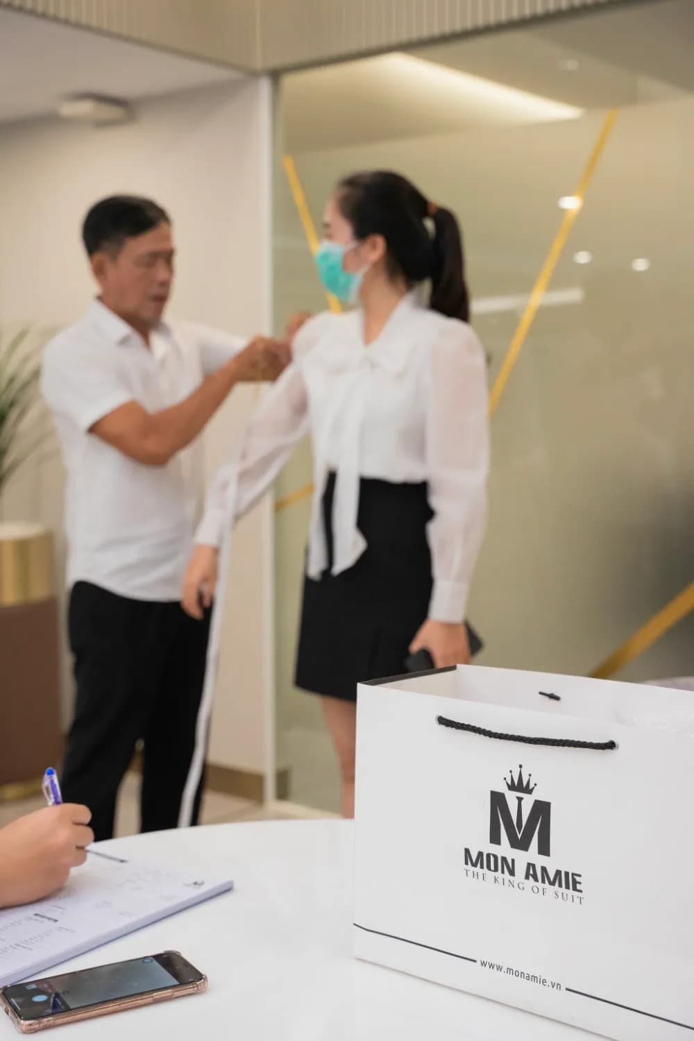 Khách nữ sơ mi trắng thắt nơ ngực được đo vai, túi giấy Mon Amie trên bàn trắng
