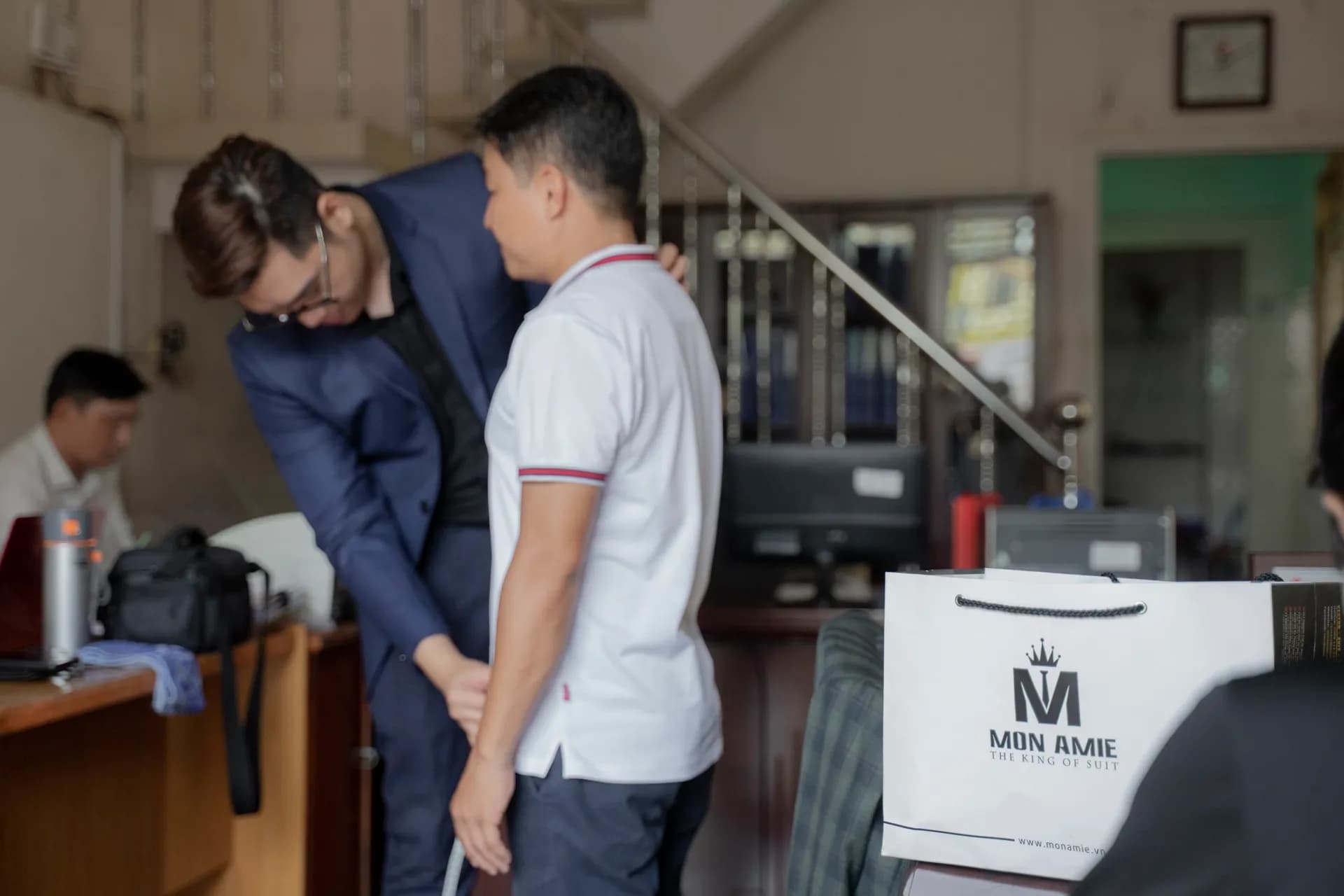 Thợ may vest navy cúi đo chiều dài chân cho khách polo trắng, túi giấy Mon Amie phía sau
